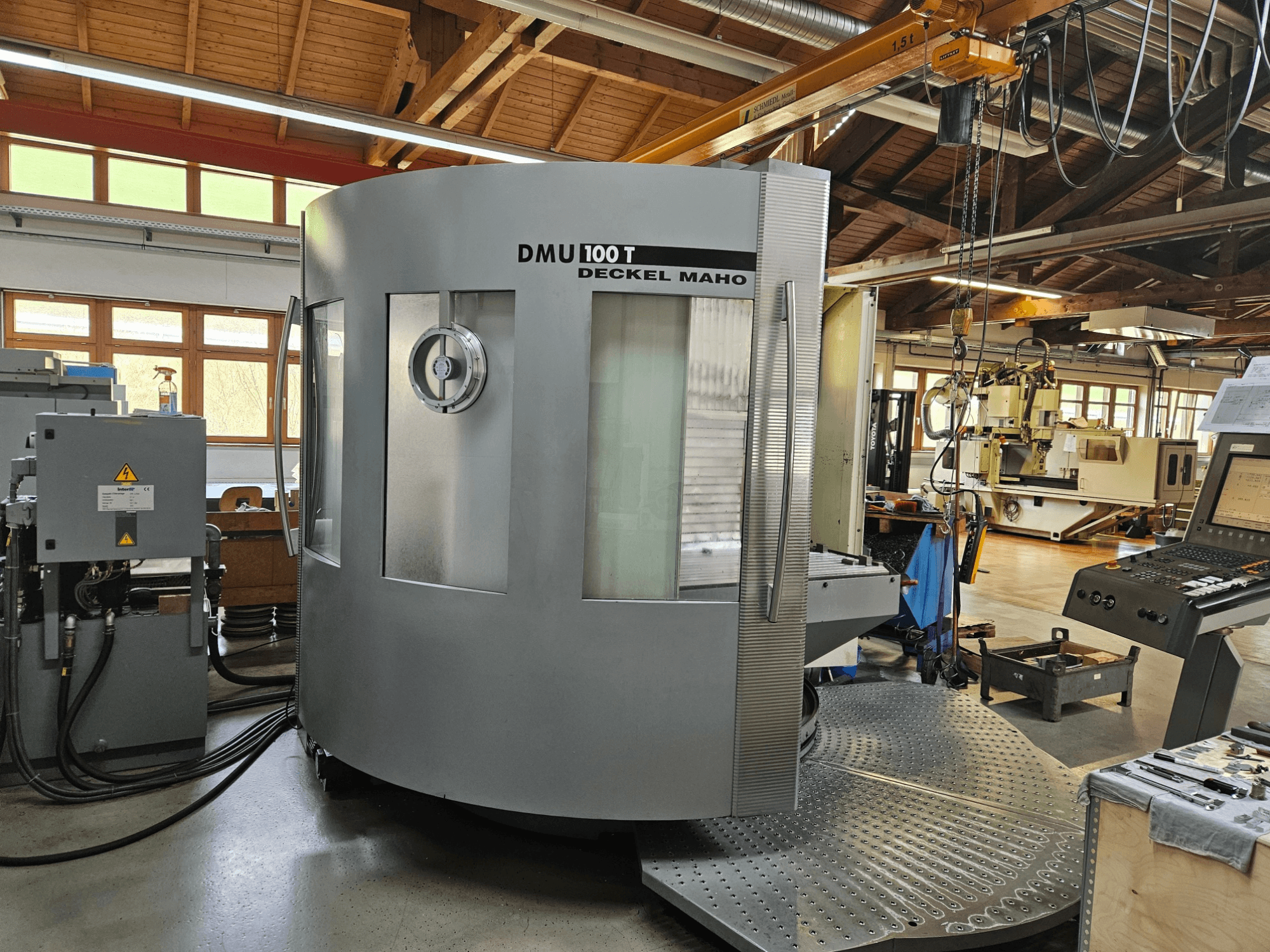 DMU 100 T Deckel Maho CNC makinesi önden bakıldığında büyük bir dairesel tabana, kontrol paneline ve cam muhafazaya sahiptir.