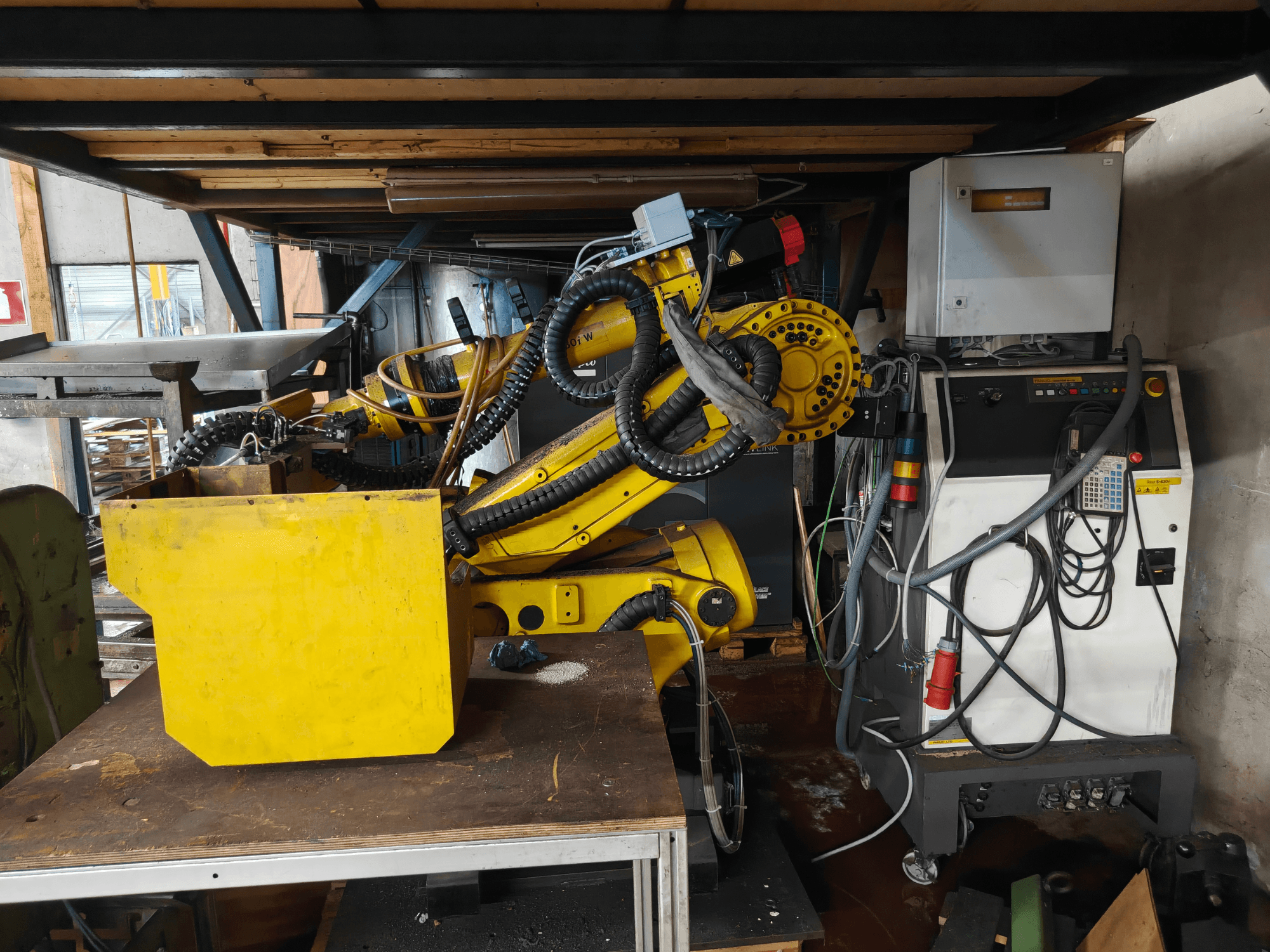 FANUC S-430i W robot kolunun önden görünümü; sarı renkli, kontrol paneli, kablolar ve çalışma tezgahı üzerinde sarı renkli bir bileşen ile birlikte.