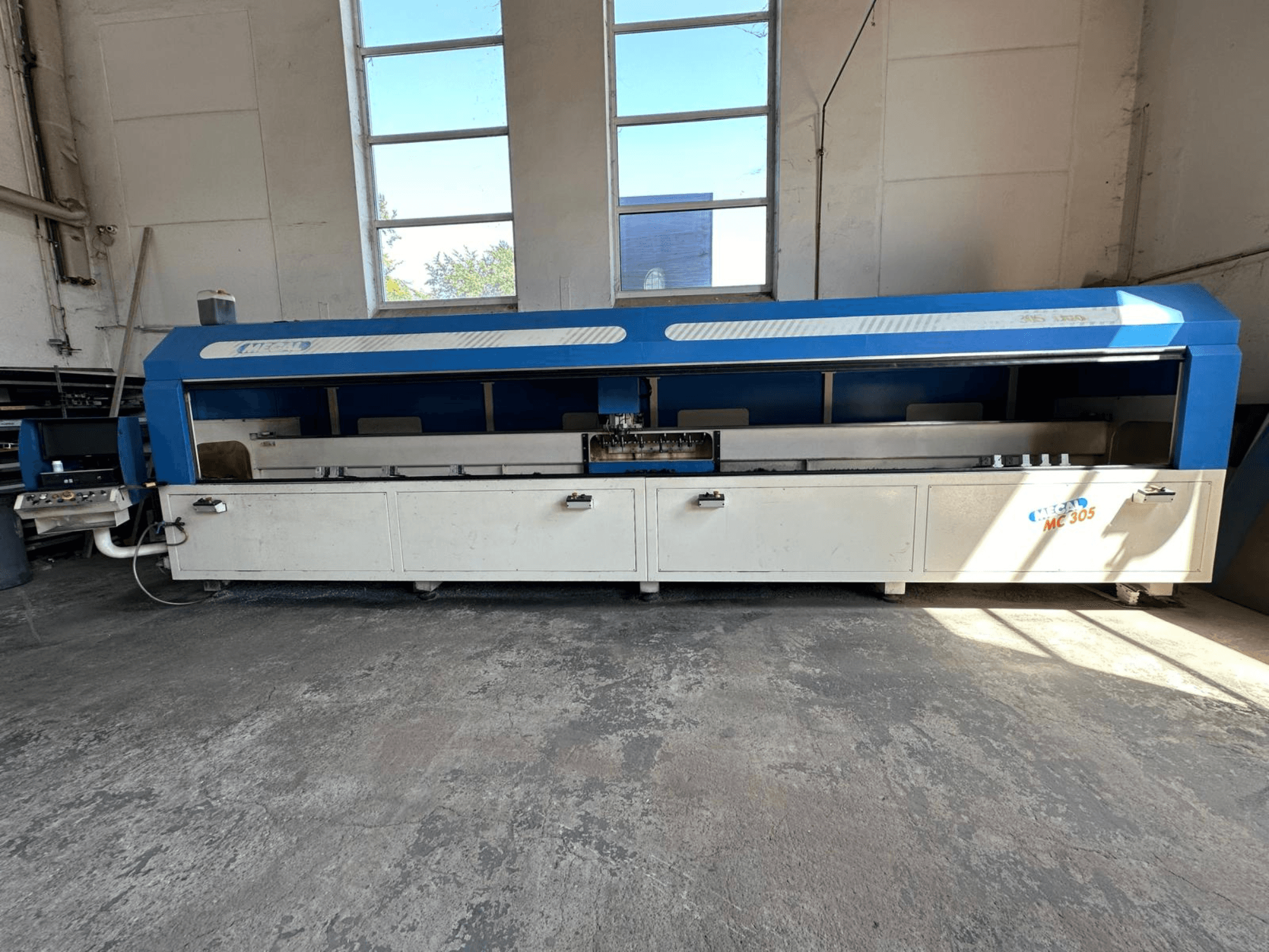 Mavi ve beyaz tasarımı, görünür çalışma masası ve kontrol paneli ile MECAL MC 305 dikey işleme merkezinin önden görünümü.