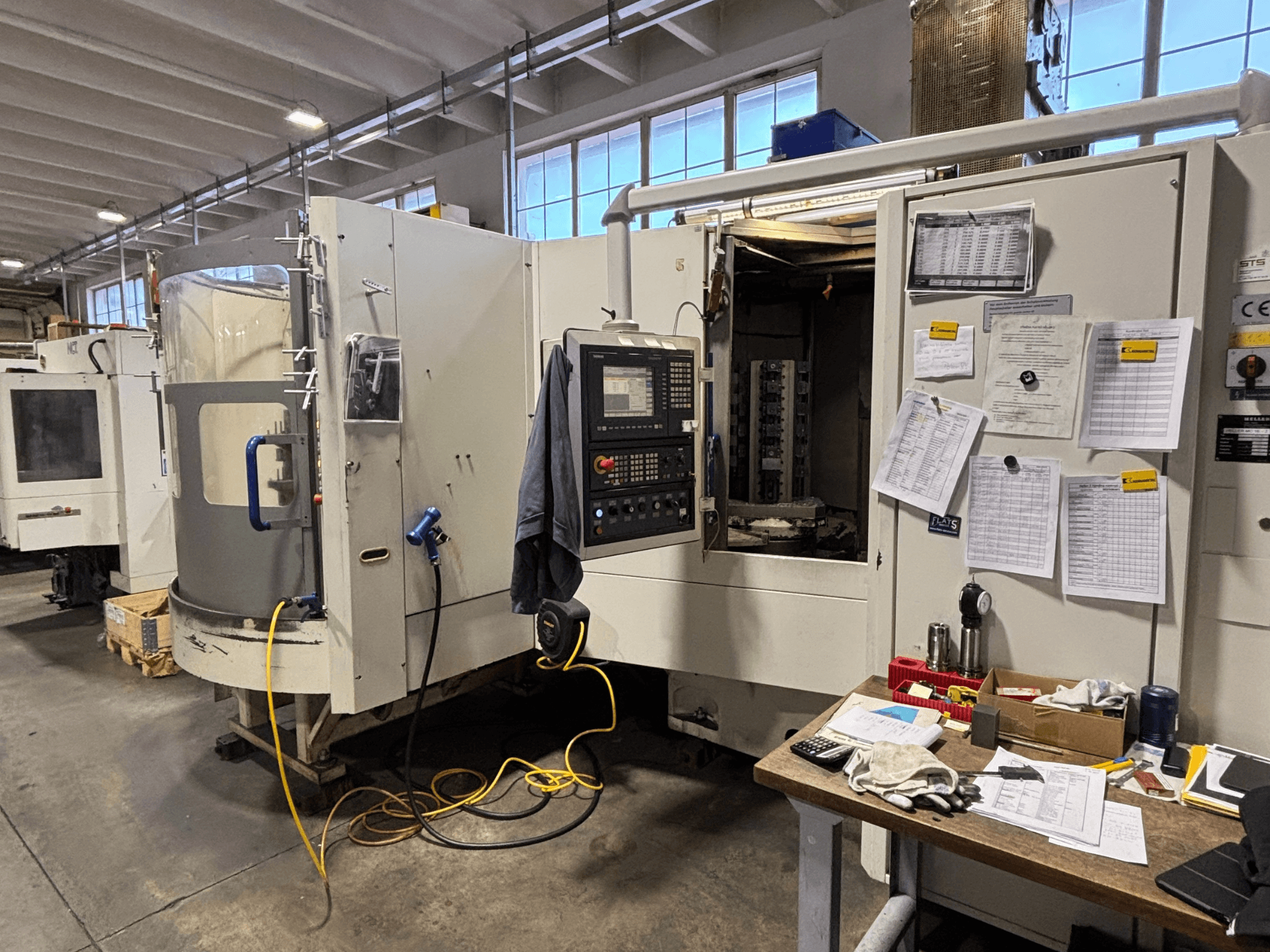 Bir atölyede CNC makinesi, önden görünüm, yan tarafta kontrol paneli ve belgeler; masada aletler ve malzemeler.