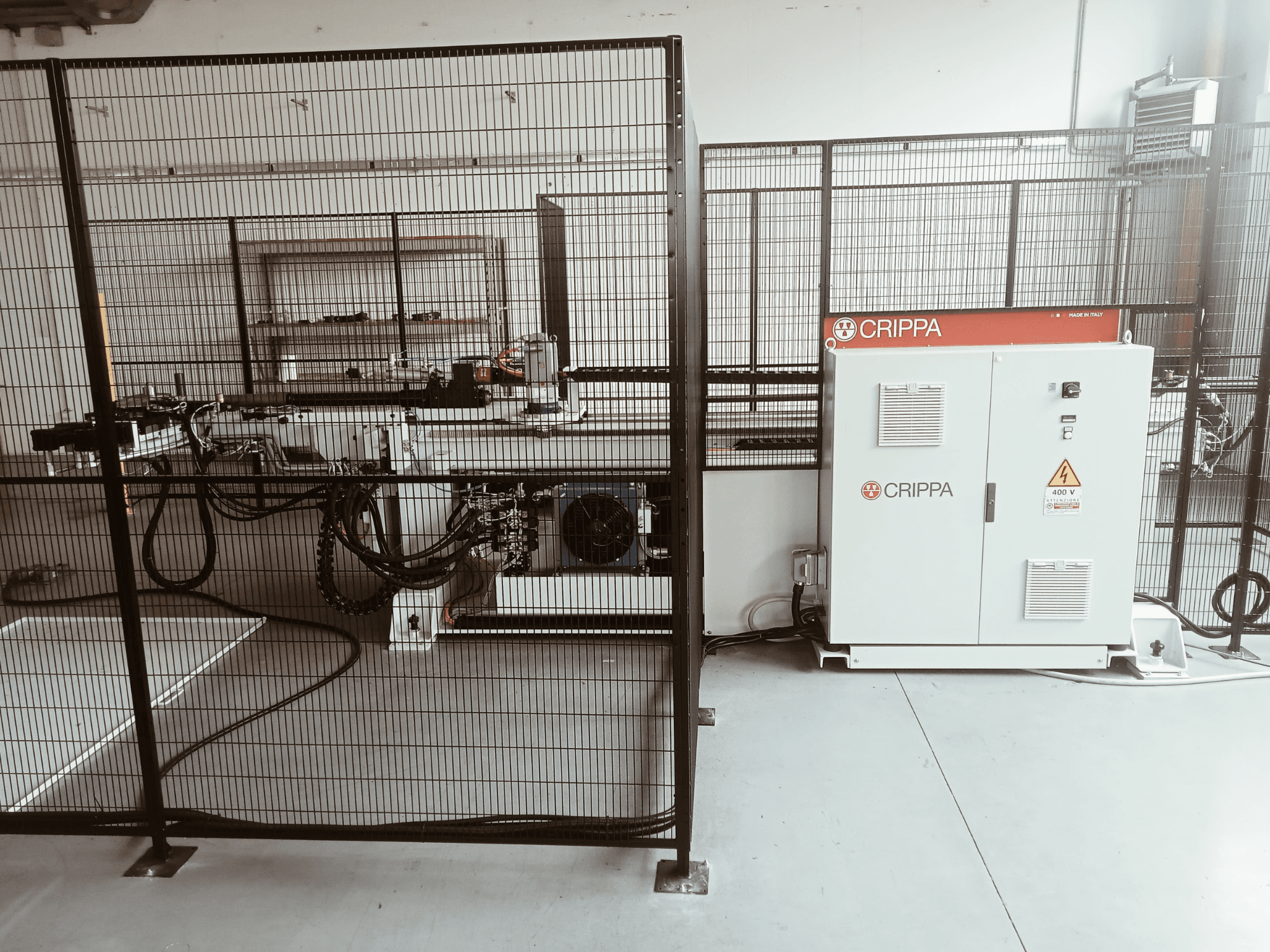 Bir güvenlik kafesinin arkasında, hortumları ve bileşenleri görünen beyaz CRIPPA makinesi, yandan görünüm, endüstriyel ortam.