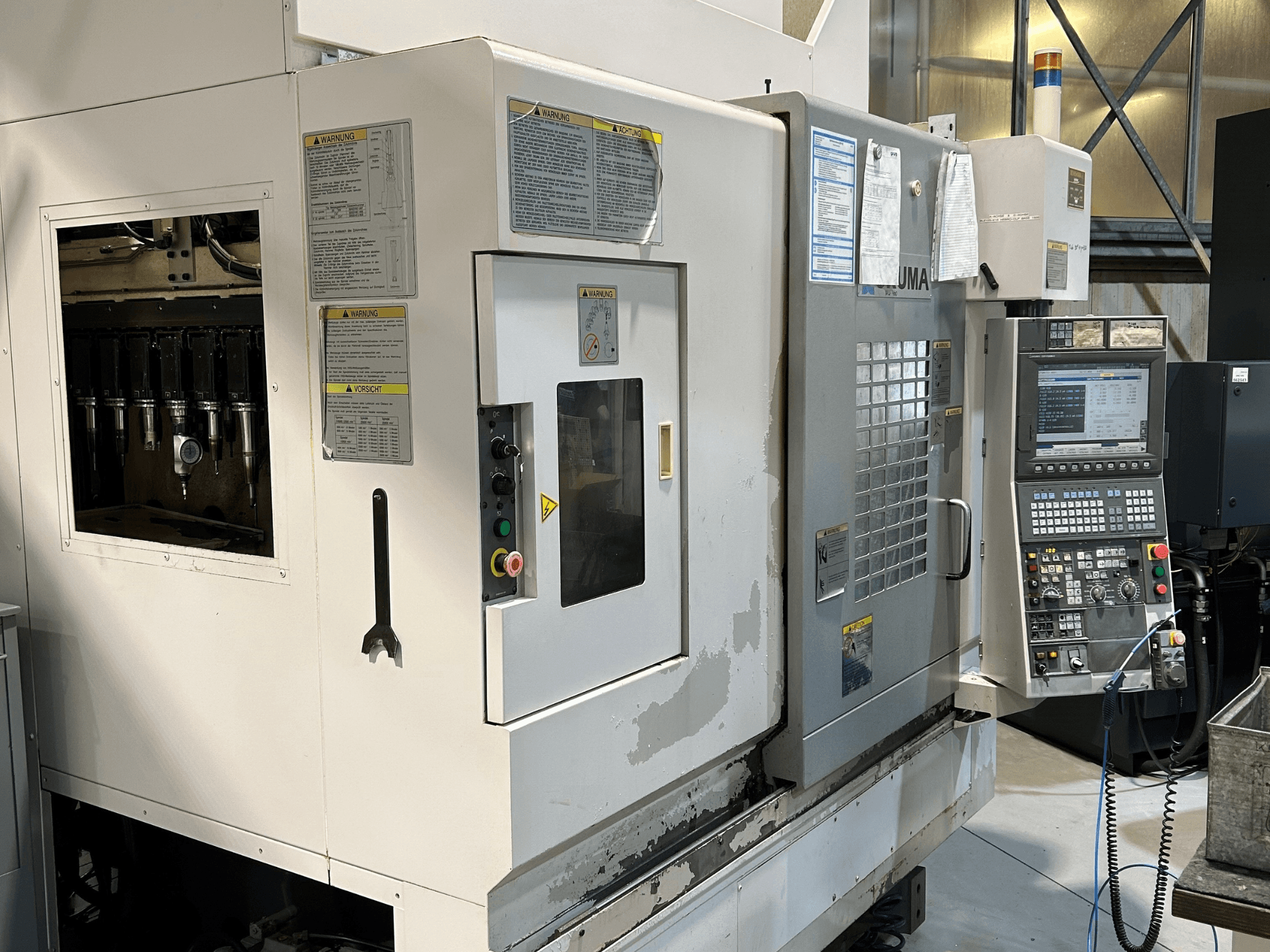 Görünür takım tutucuları, kontrol paneli, uyarı etiketleri ve çalıştırma düğmeleri ile bir UMA CNC makinesinin önden görünümü.