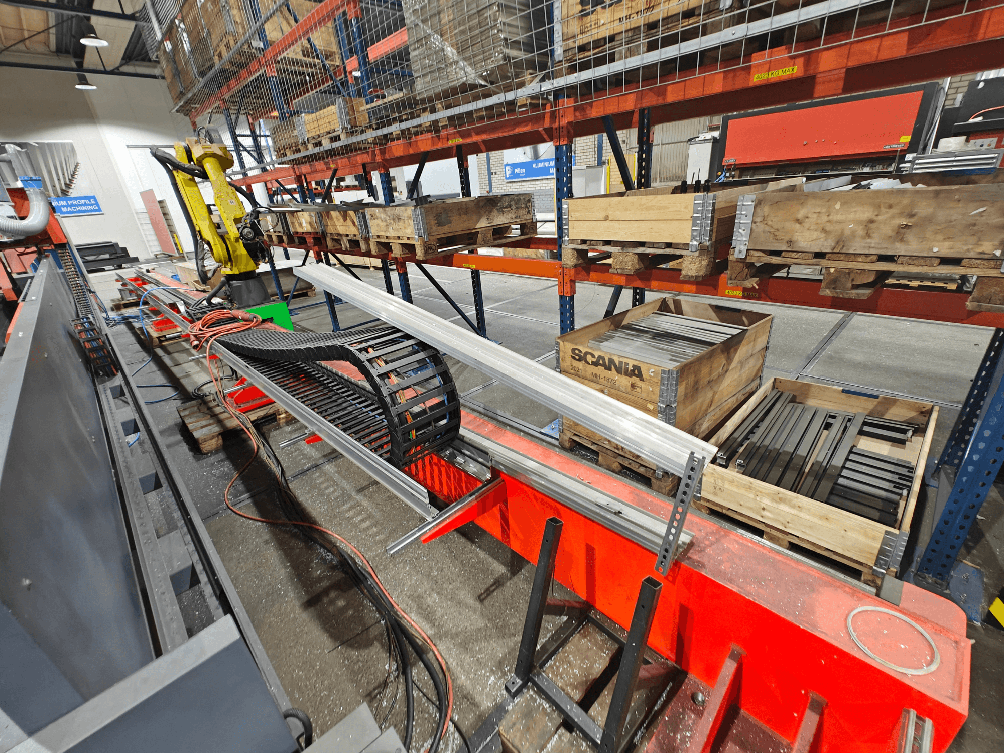 İş istasyonu üzerindeki robotik kol, turuncu ve siyah kablolar görünür, Scania kasaları ve alüminyum profiller fabrika ortamında.