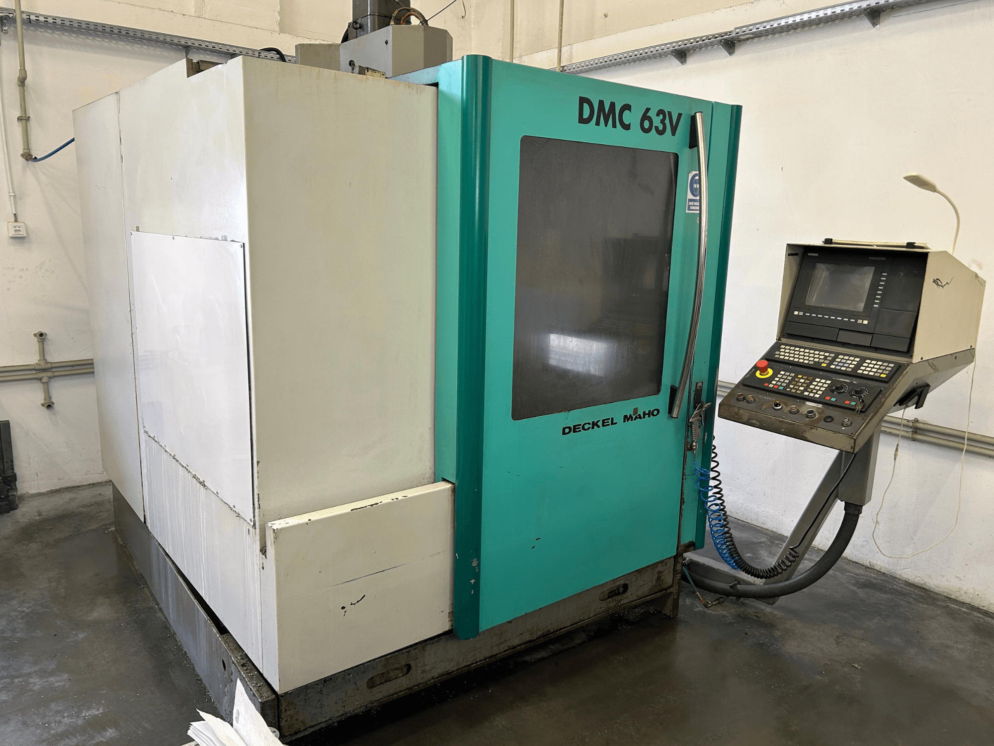 Dikey işleme merkezi DECKEL MAHO DMC 63V'nin önden görünümü, kontrol paneli ve mavi-yeşil kasası.