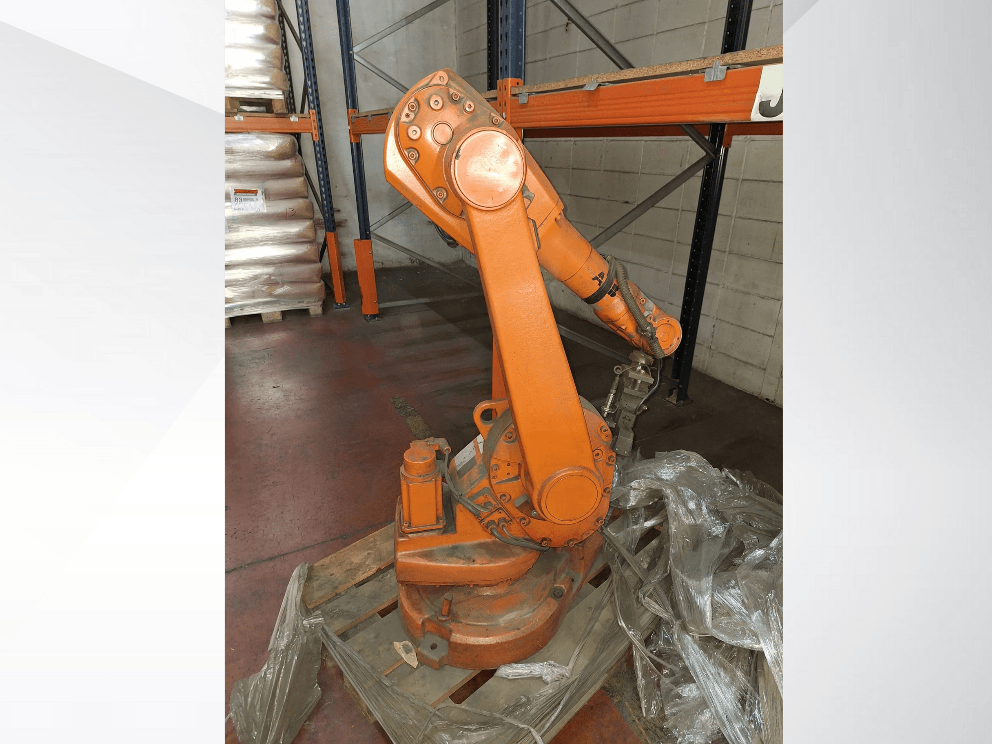 Turuncu renkli endüstriyel robotik kol, ahşap palet üzerinde önden görünüm, bağlı kablo ve taban etrafında koruyucu plastik sargı.