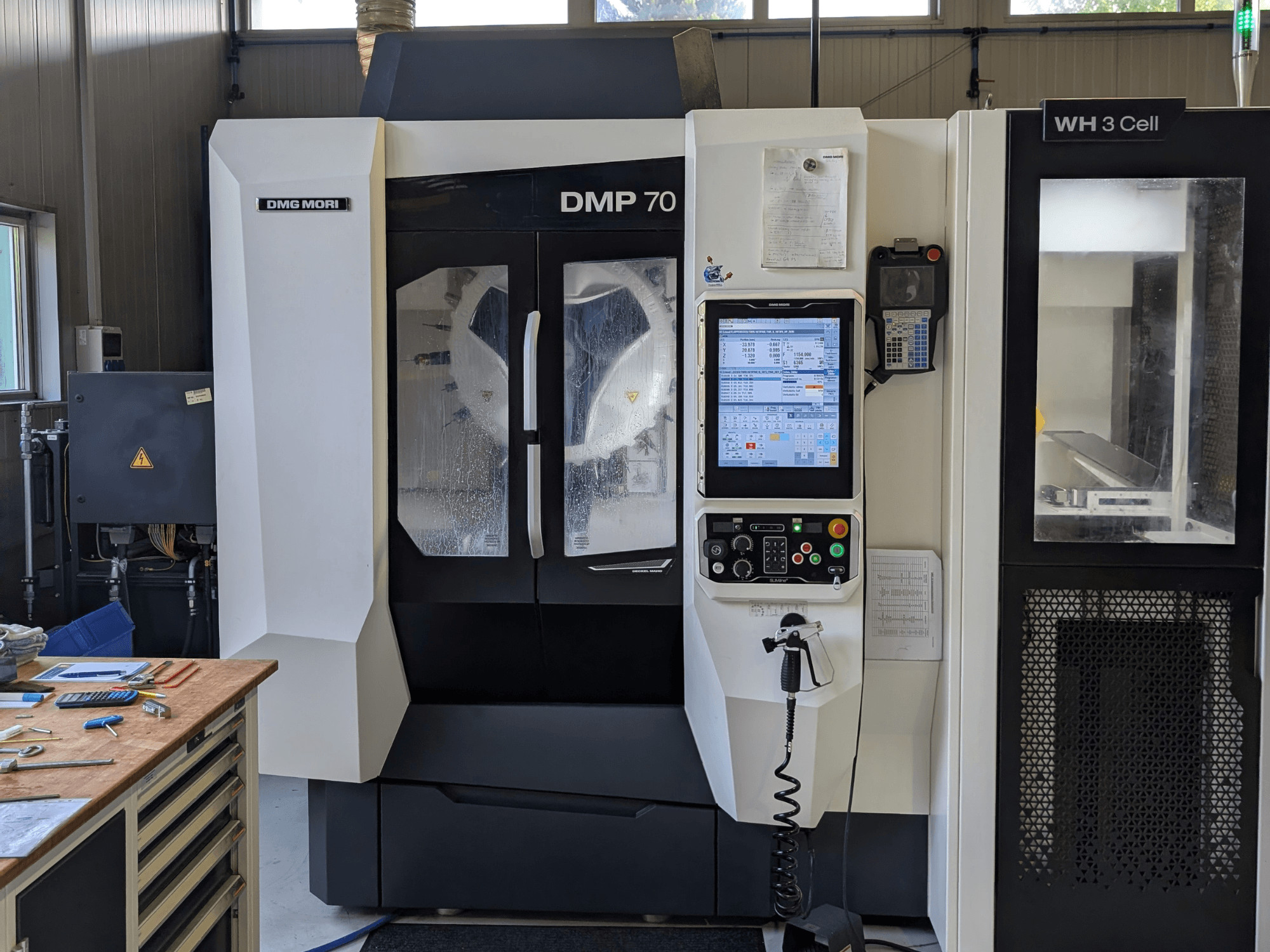 DMG MORI DMP 70 dikey işleme merkezi; kontrol paneli ve çalışma ekranı ile birlikte önden görünüm, etrafında atölye aletleri yer almaktadır.