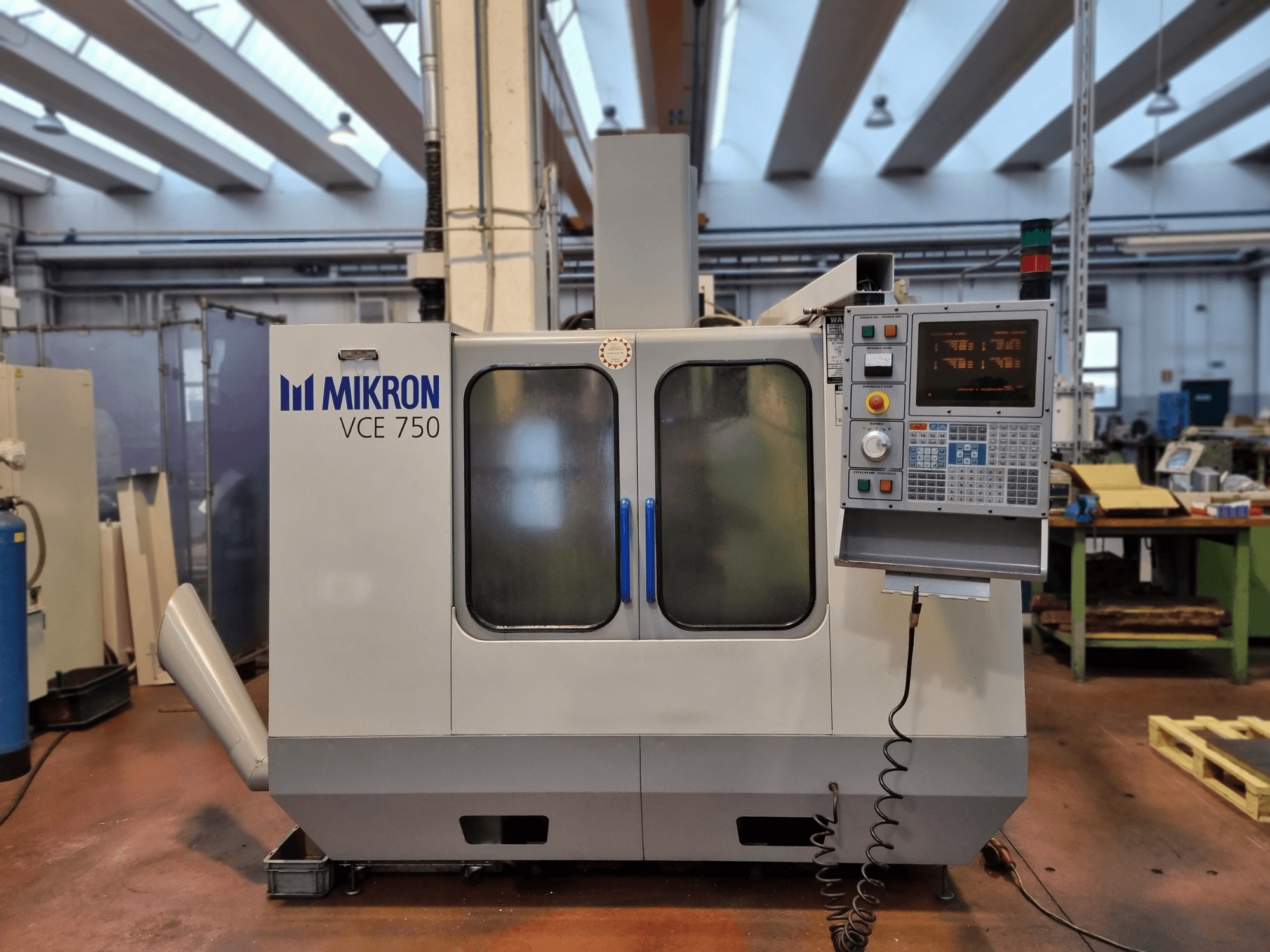 Bir atölyede beyaz Mikron VCE 750 CNC makinesi, yandan görünüm, dijital kontrol paneli ve büyük cam kapılar.
