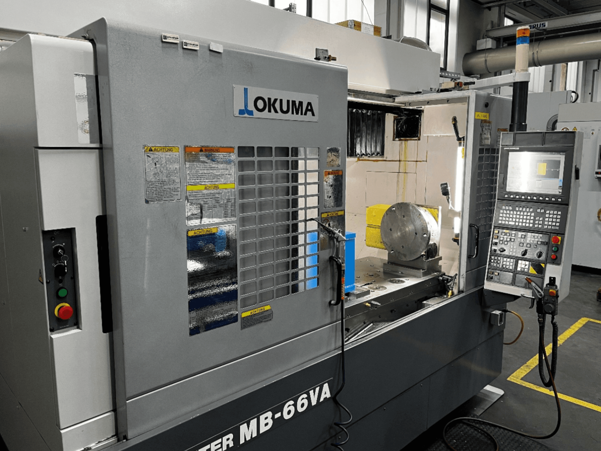 Okuma MB66-VA Dik İşleme Merkezi kontrol paneli, iş parçası ve güvenlik etiketleri ile makine içini gösteren önden görünüm.