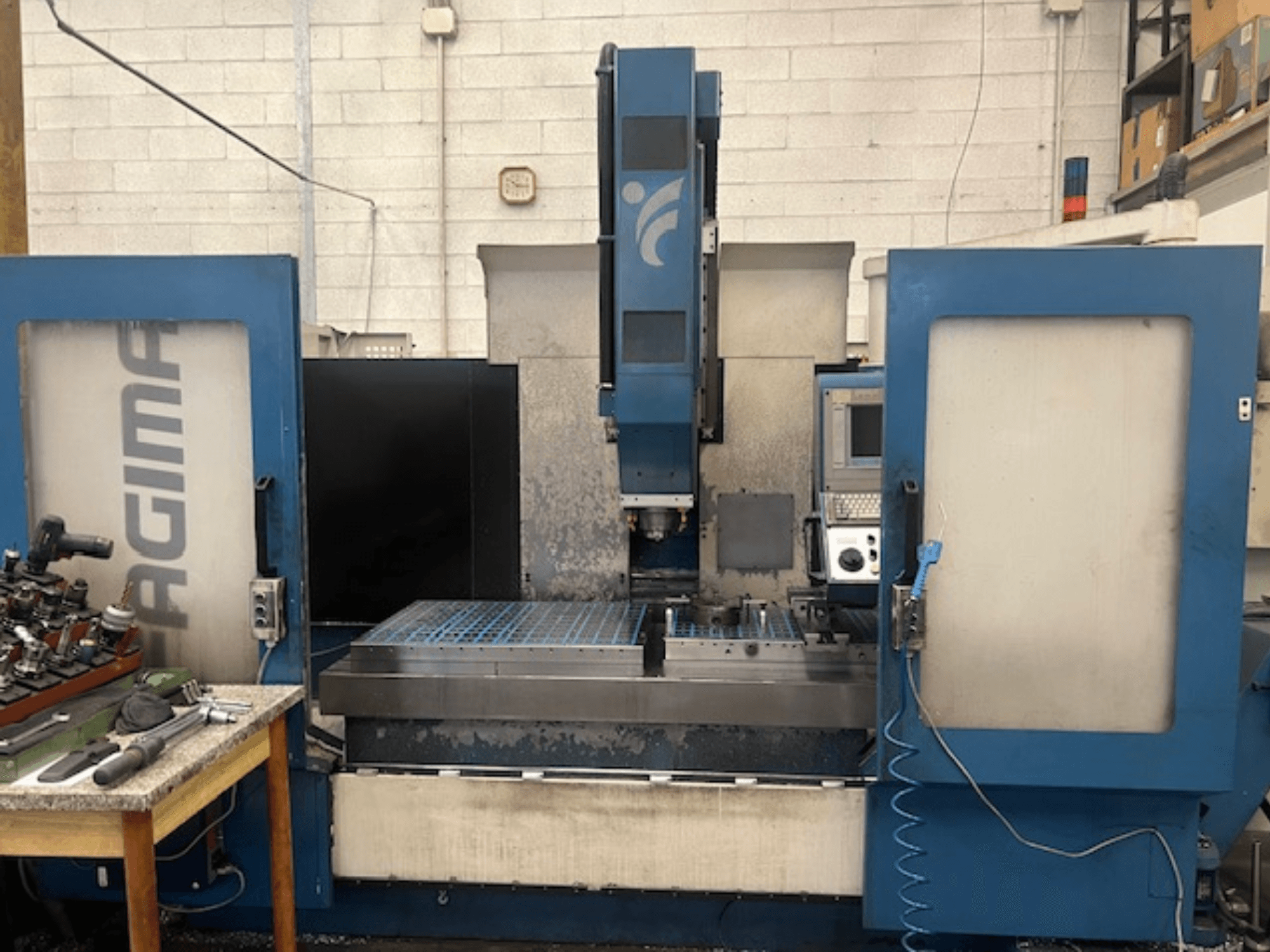 Büyük bir metal çalışma alanı, görünür kontrol paneli ve yakındaki bir masada takımları olan bir JAGIMA CNC makinesinin önden görünümü.