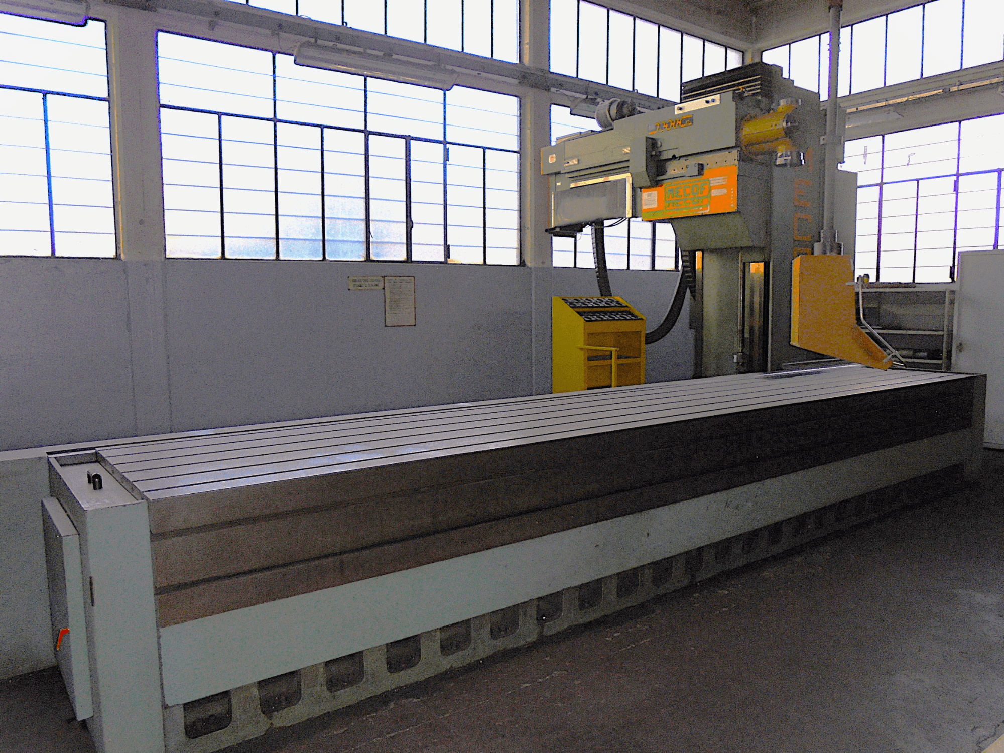 Markası bilinmeyen, bir fabrikadaki dikey freze makinesi, yandan görünüm, yivli yüzeye ve kontrol paneline sahip büyük bir tablaya sahiptir.