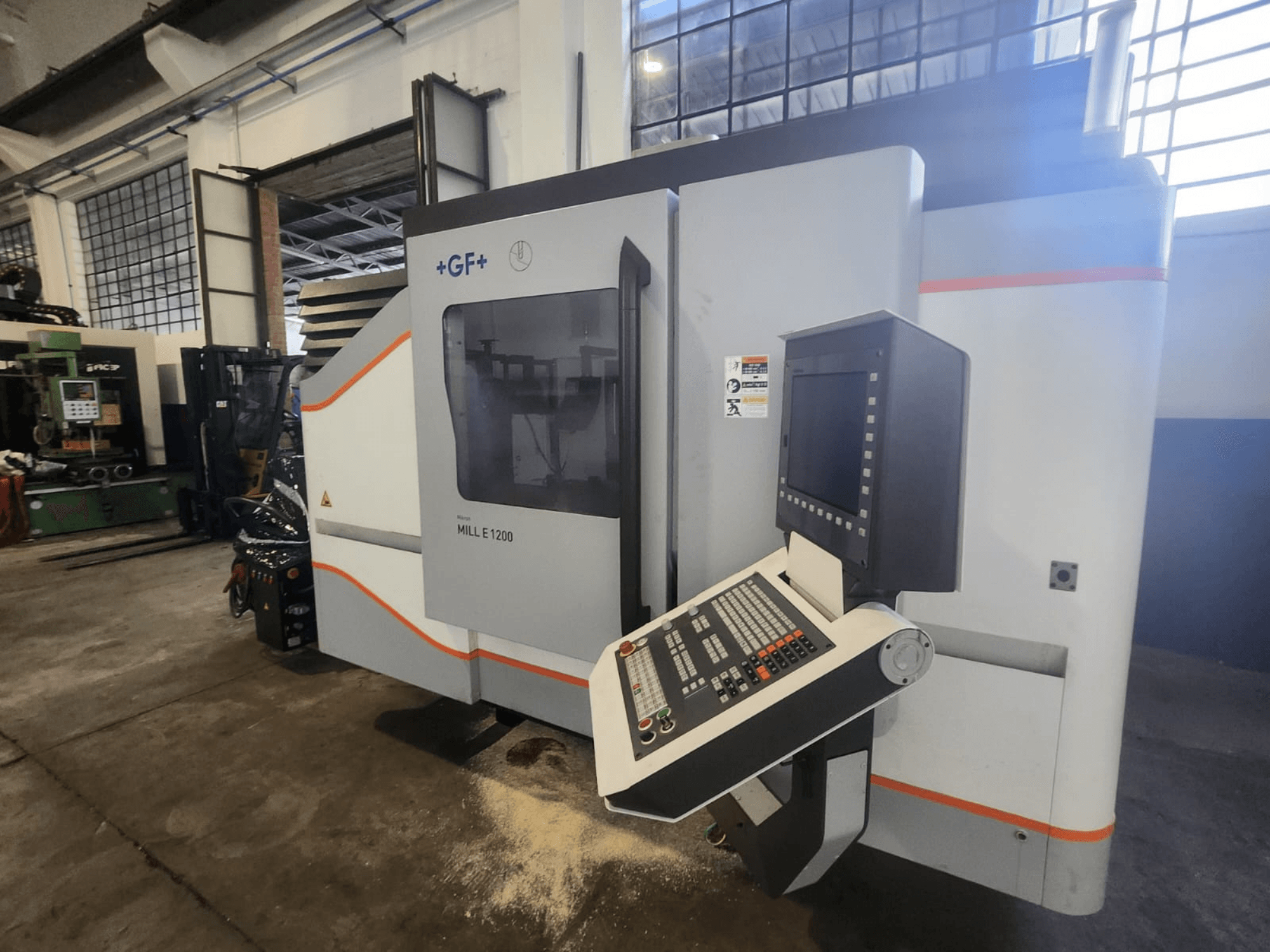 GF MILL E1200, atölye ortamında kontrol paneli, ekran ve güvenlik etiketleri ile freze makinesinin önden görünümü.