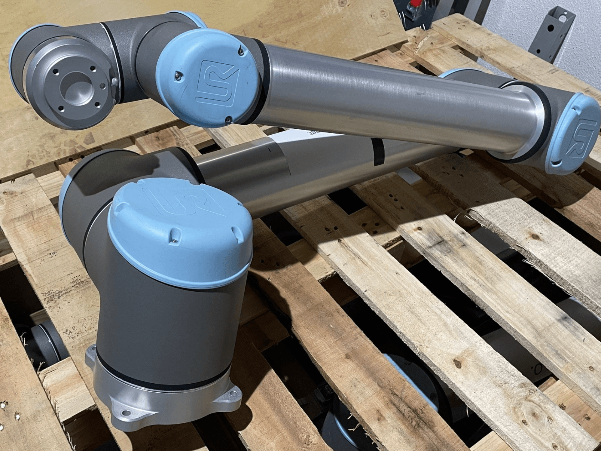 Mavi uç kapaklı gümüş robotik kol, önden görünüm, eklemler ve mekanik detaylar sergileniyor, ahşap bir palet üzerinde duruyor.