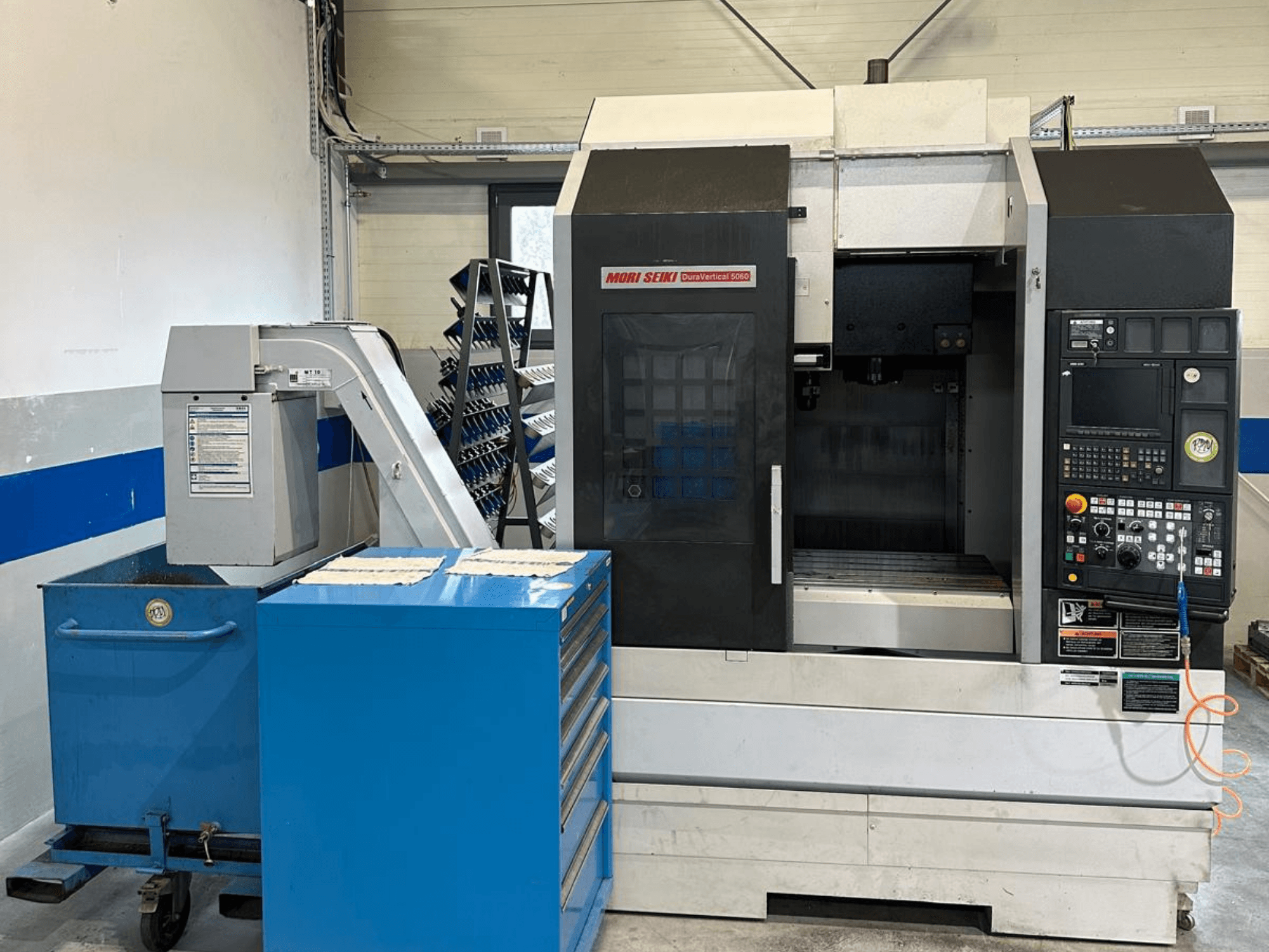 MORI SEIKI DURA VERTICAL 5060, kontrol paneli ve yanındaki takım deposu ile birlikte bir dikey işleme merkezinin önden görünümü.