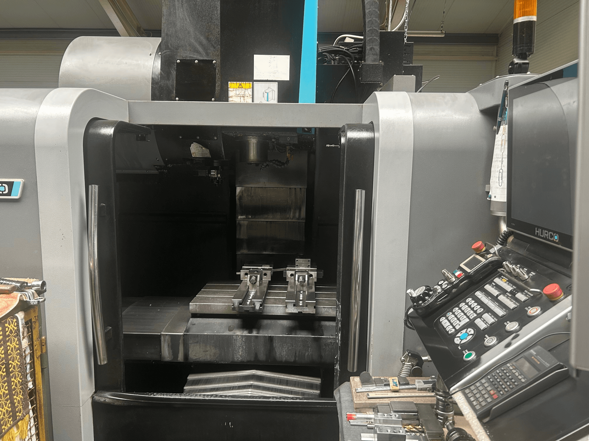 Hurco CNC işleme merkezi önden görünüm, takım tutucu, metal iş parçası ve düğmeli ve ekranlı kontrol panelini gösterir.