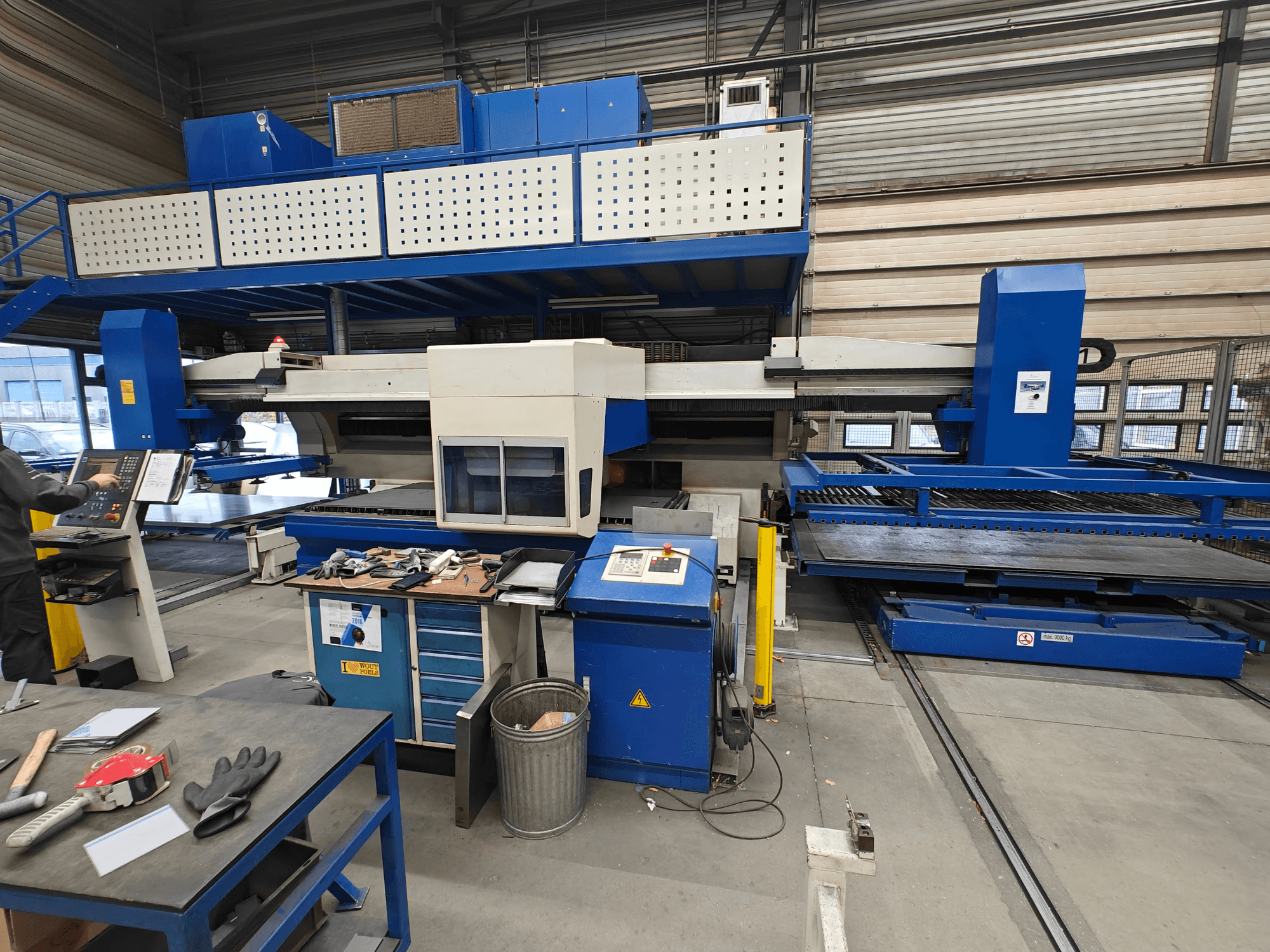 Önden görünümlü endüstriyel CNC makinesi, mavi ve beyaz yapı, kontrol paneli ve çalışma masası üzerinde çeşitli aletler.