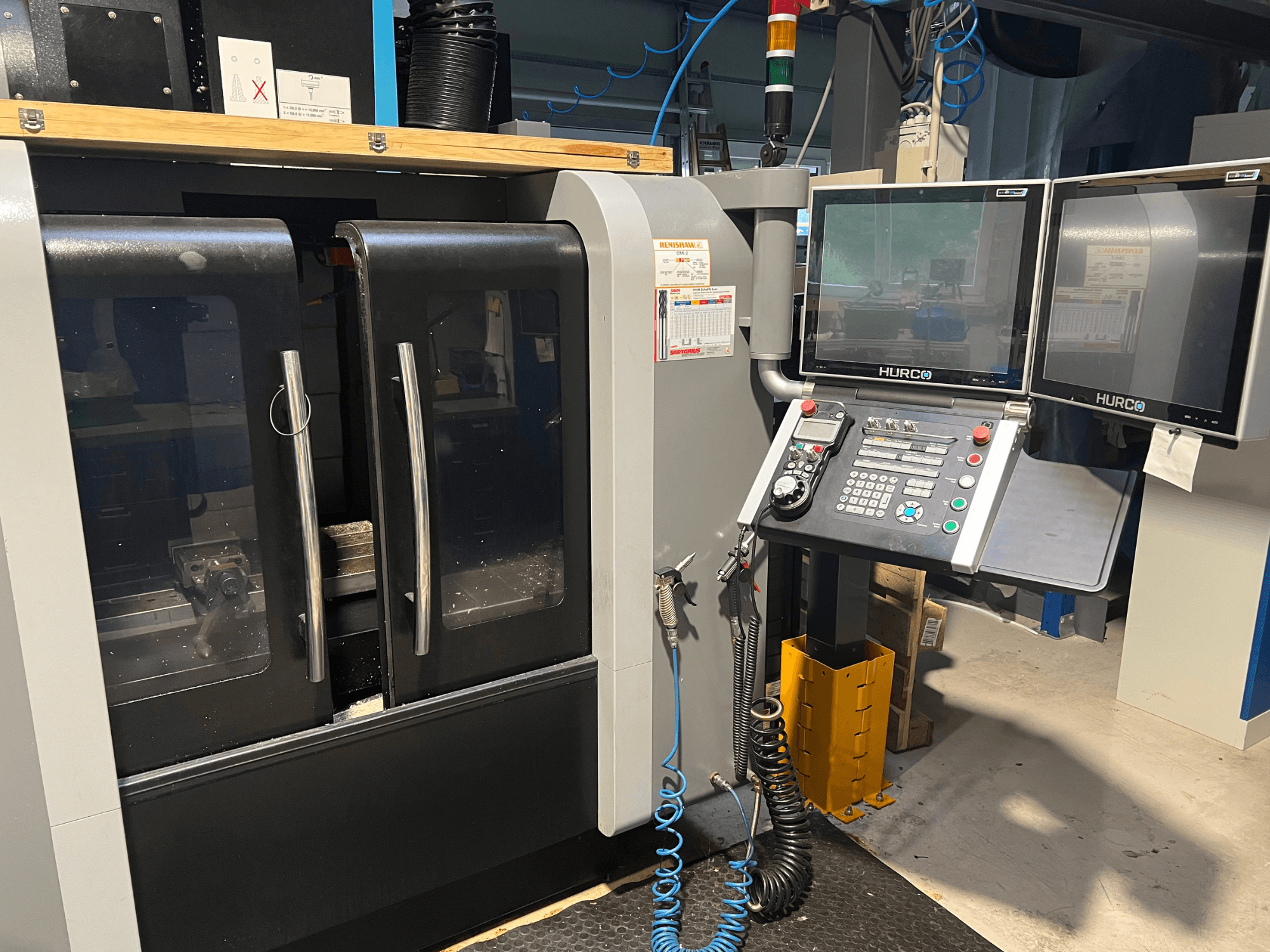 Çift monitörlü ve kontrol panelli Hurco CNC makinesinin önden görünümü. Şeffaf kapılar ve içinde görünür işleme alanı bulunur.