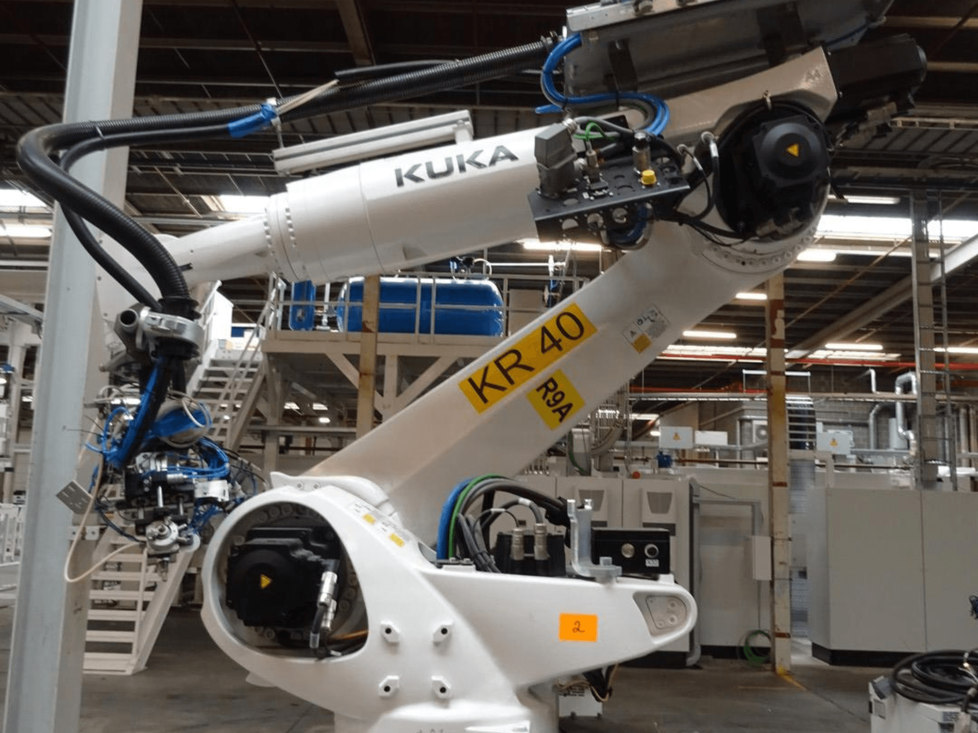 KUKA KR 120 R3100-2 robot kolu yandan görünümde, kontrol paneli ve mekanik bileşenleri sergiliyor, endüstriyel bir ortama yerleştirilmiş.