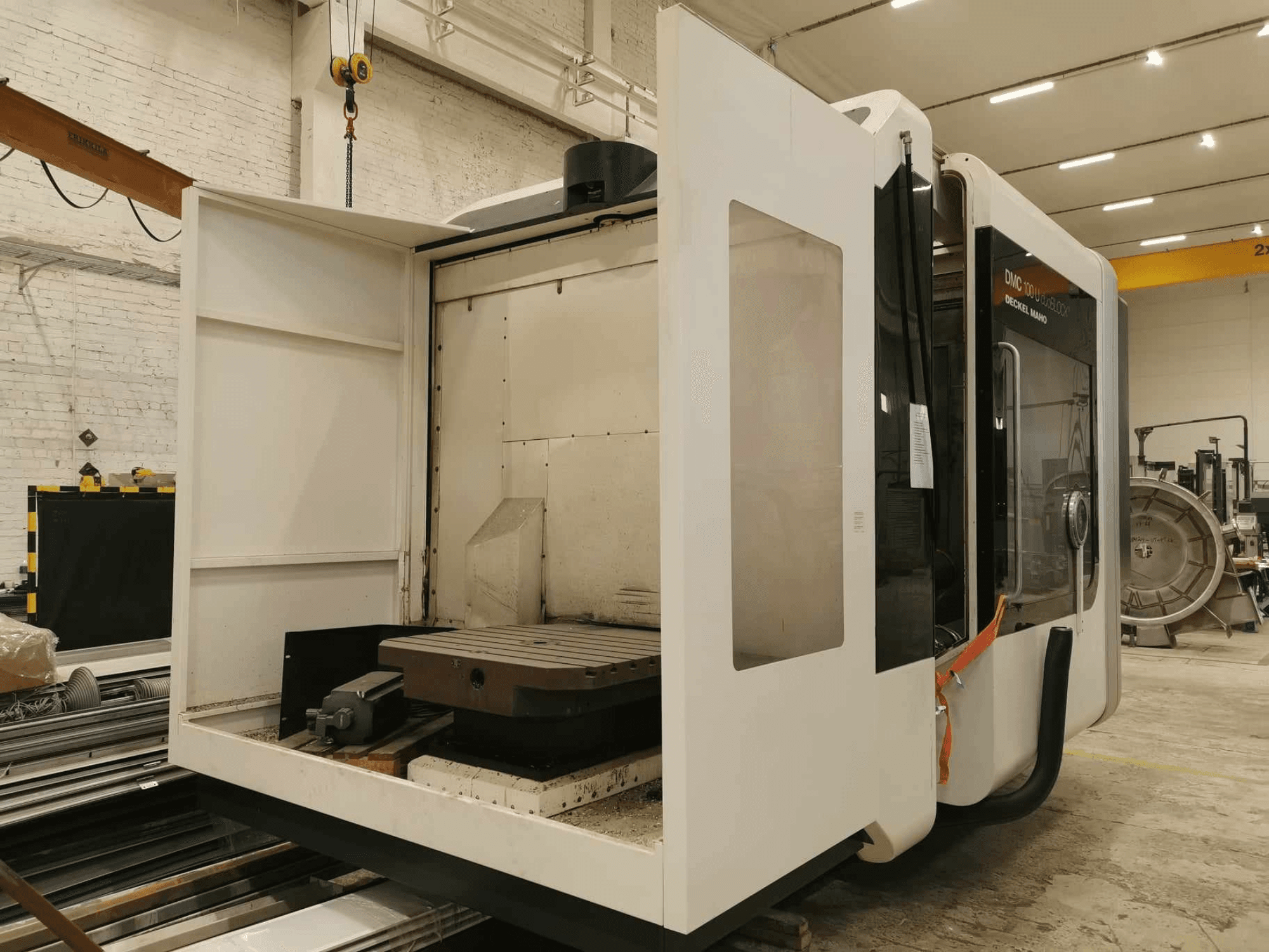 DMC 100U lineer CNC makinesi, açık erişim kapısı ile yandan görünüm, çalışma masası ve dahili bileşenleri gösterir.