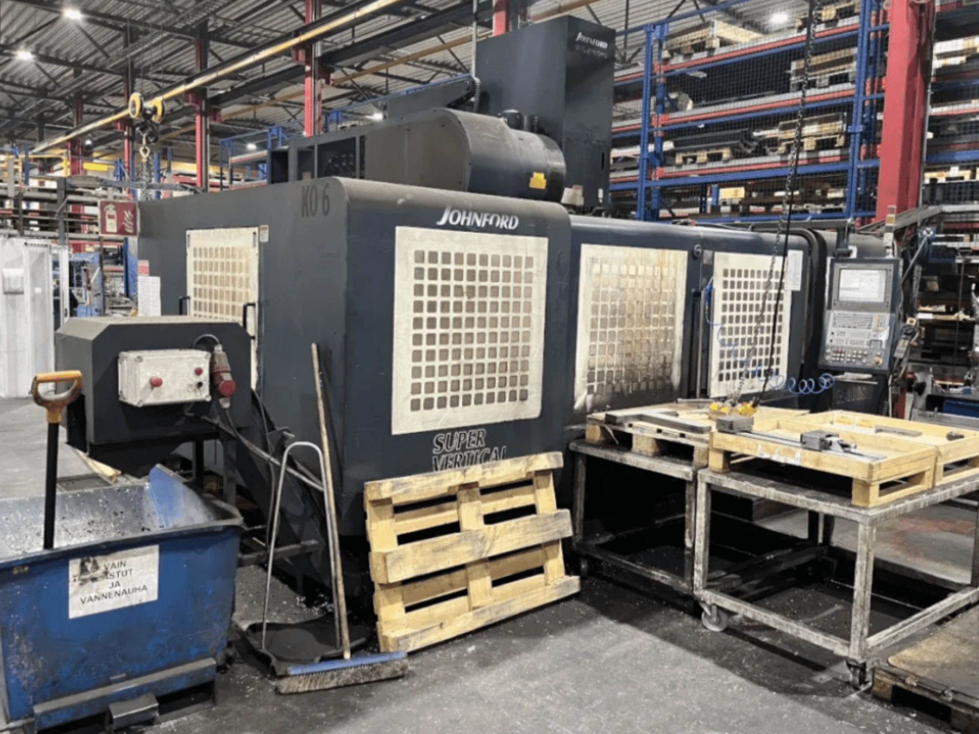 Fabrikadaki Johnford Süper Dikey freze makinesi, kontrol paneli, paletler ve yakındaki temizlik ekipmanı ile yandan görünüm.