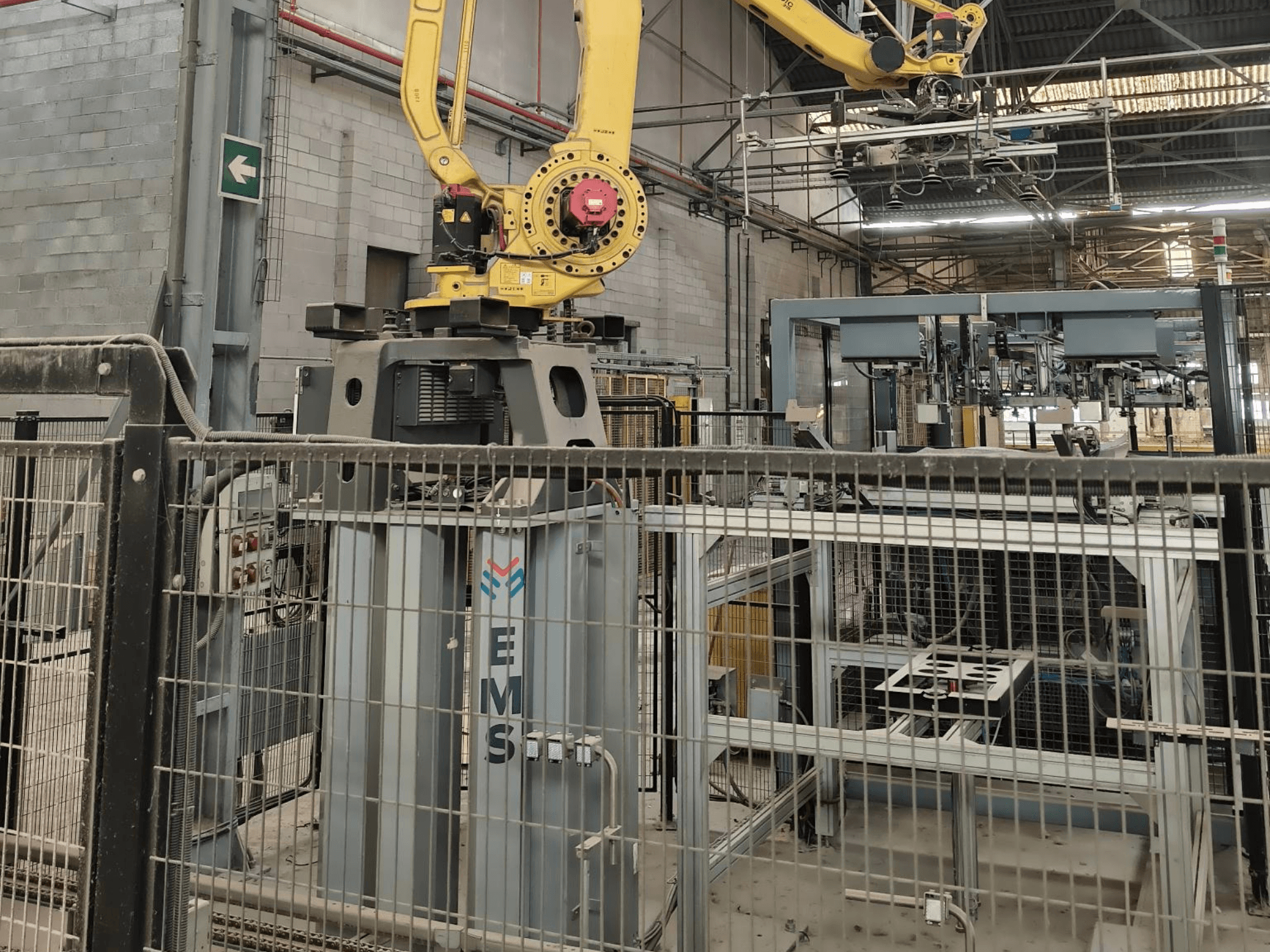 Arka planda endüstriyel ekipmanlar ve koruma çitleri bulunan, bir EMS aletinin üzerine yerleştirilmiş FANUC M-410iC/315 robot kolunun önden görünümü.