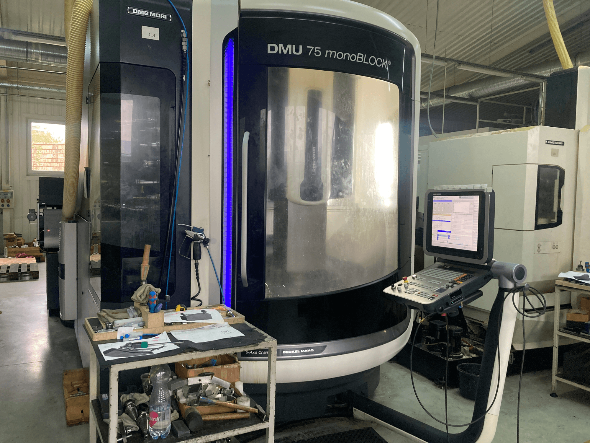 DMG MORI DMU 75 monoBLOCK® CNC makinesi, önden görünüm, kontrol paneli, aletler ve malzemelerin bulunduğu bir çalışma alanı.