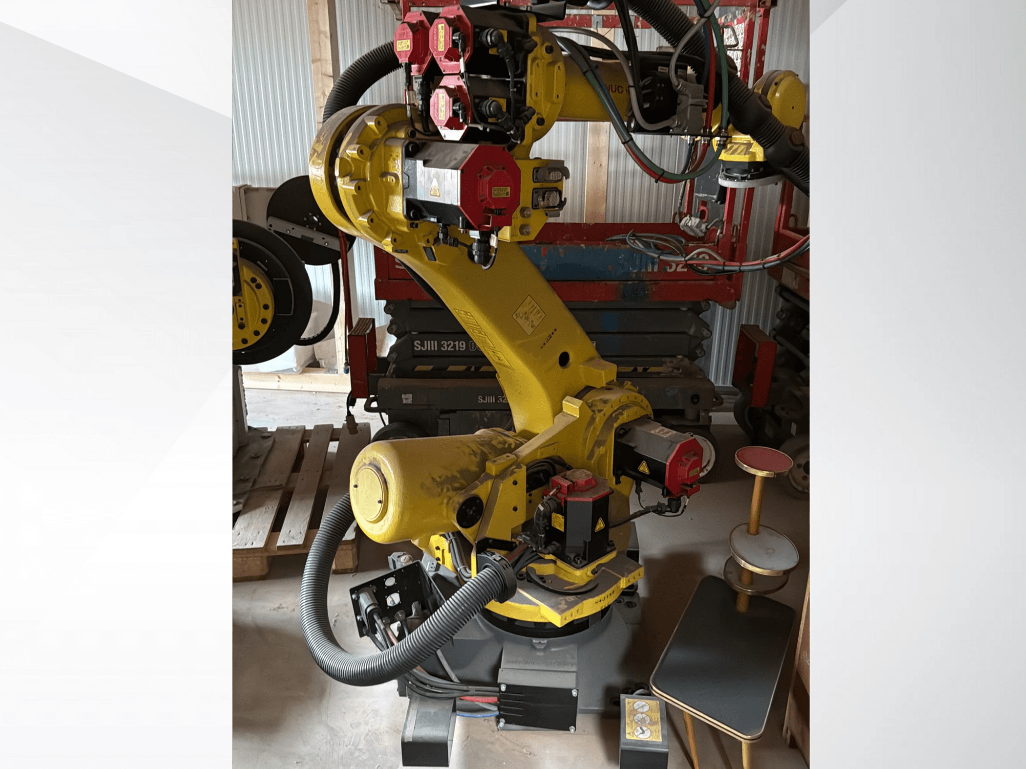 Kırmızı kontrolörlü sarı endüstriyel robotik kol, önden görünüm; çoklu eklem ve hortumlara sahiptir, bir çalışma sahasına yerleştirilmiştir.