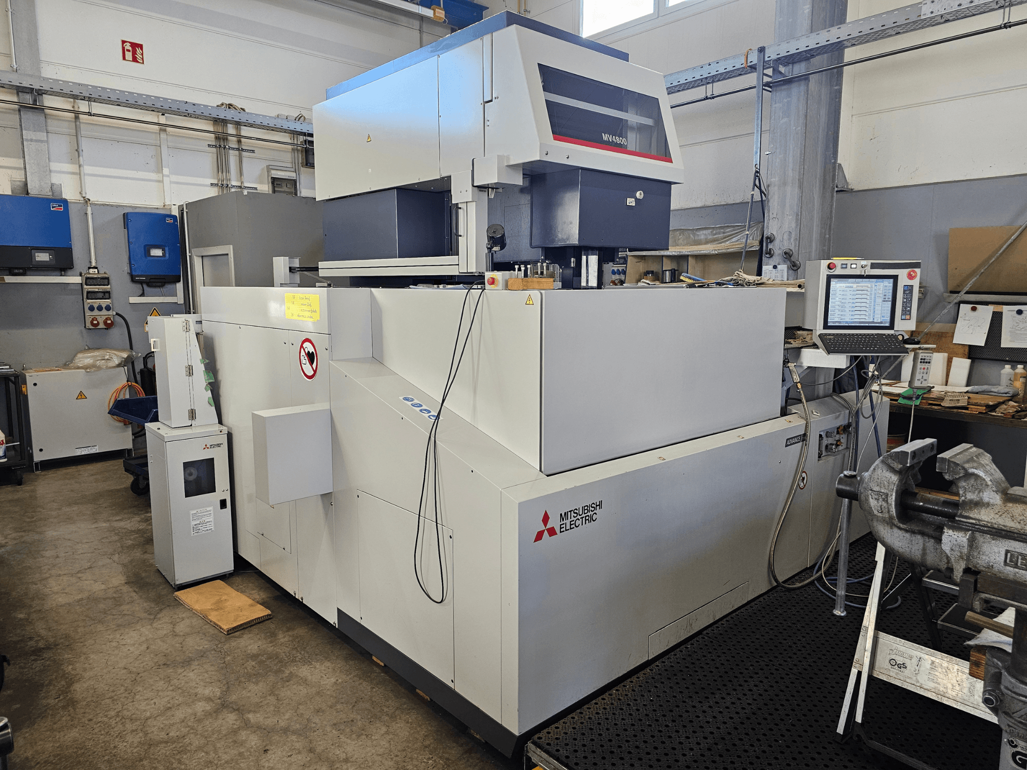 Bir atölyedeki Mitsubishi MV4800 Tel Erozyon Makinesi, kontrol panelini, güvenlik etiketlerini ve çevresindeki ekipmanı gösteren yandan görünüm.