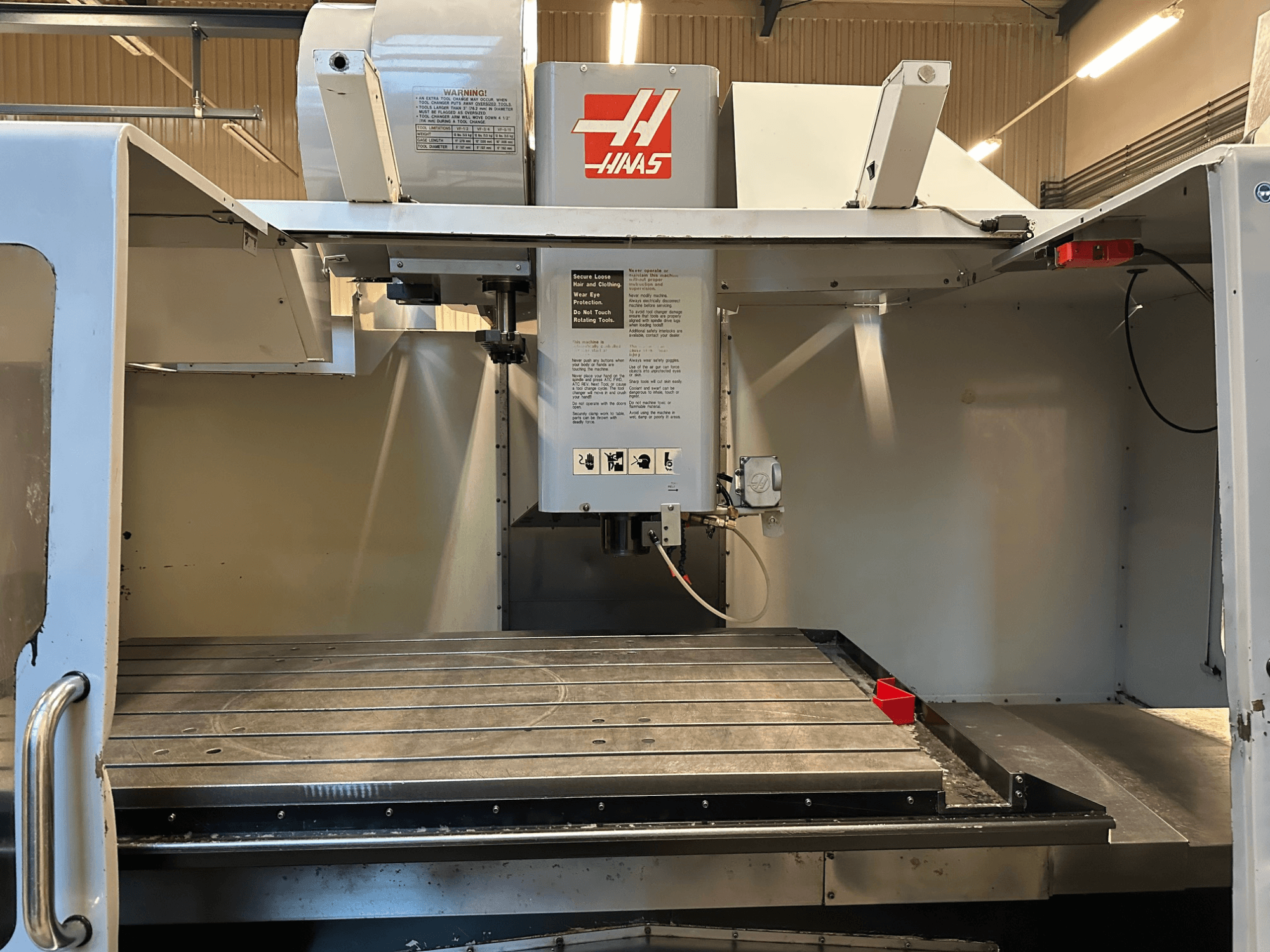 Haas CNC freze makinesi, model VF-2, kontrolleri gösteren önden görünüm, metal çalışma masası ve panel üzerindeki güvenlik etiketi.