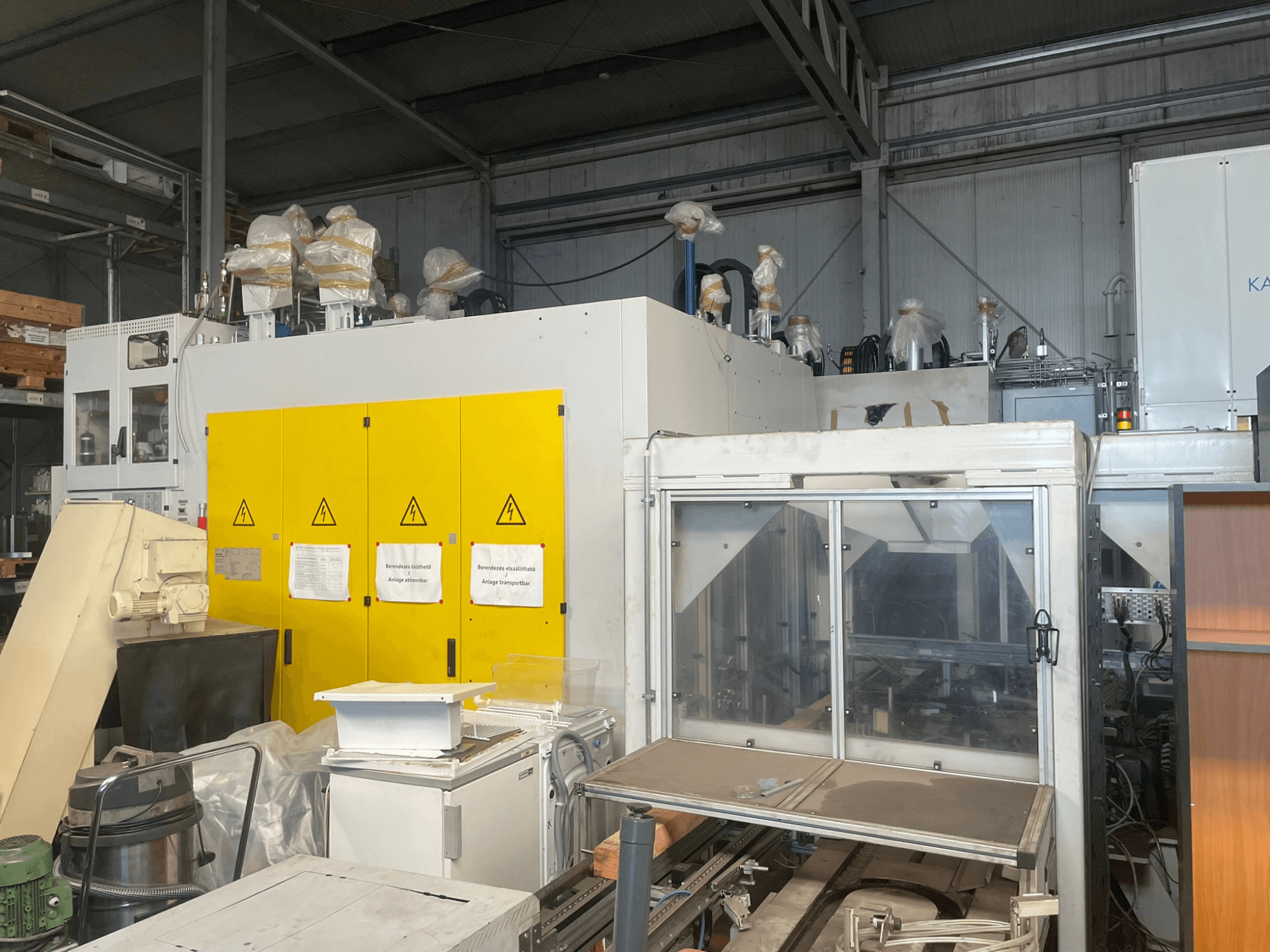 Depo ortamında, sarı kontrol panelli, yandan görünümlü, uyarı etiketli ve plastik kaplamalı endüstriyel makine.