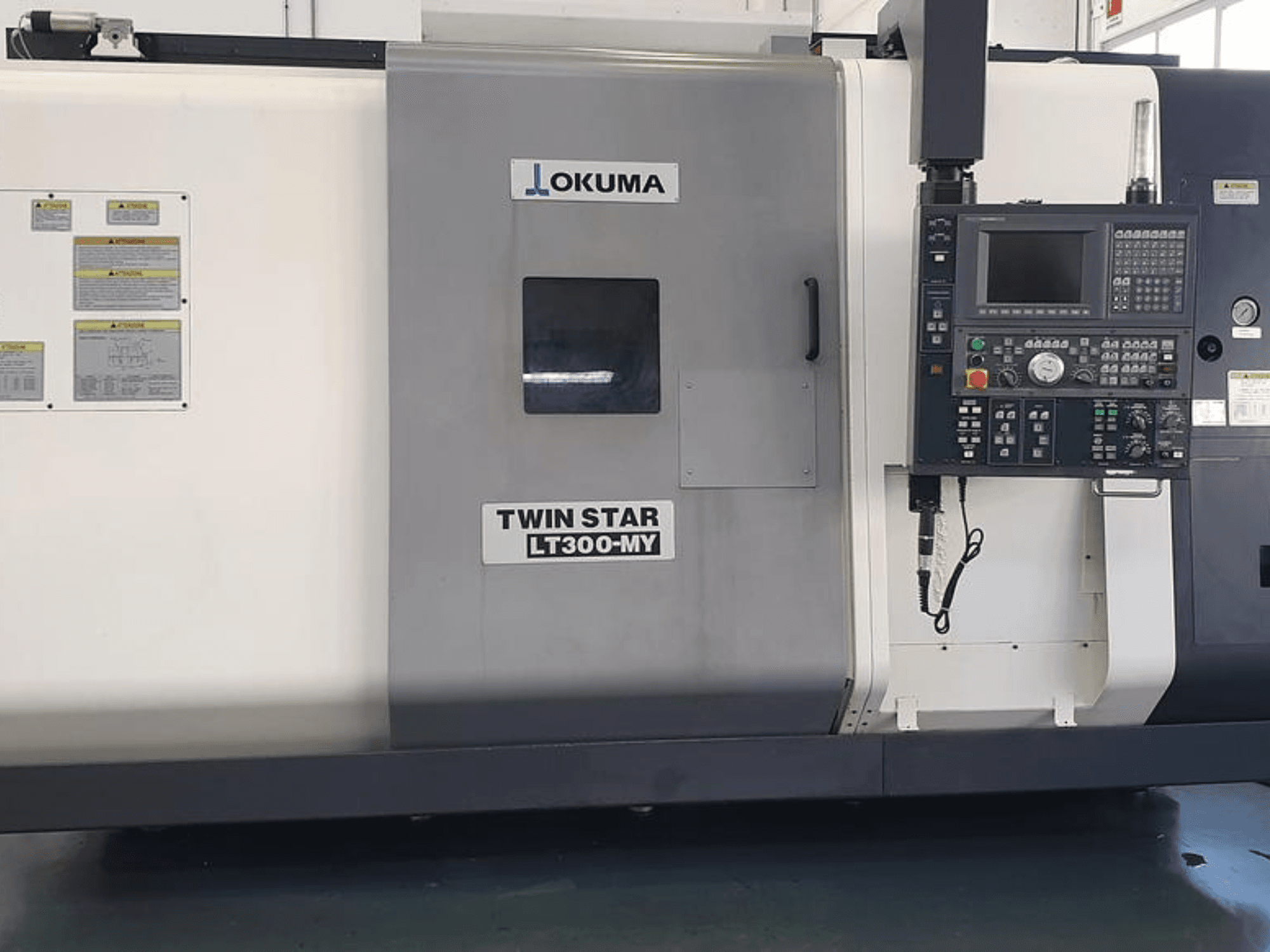 Okuma Twin Star LT300-MY CNC torna tezgahının yandan görünümü, kontrol paneli, çalışma düğmeleri ve güvenlik etiketleri.