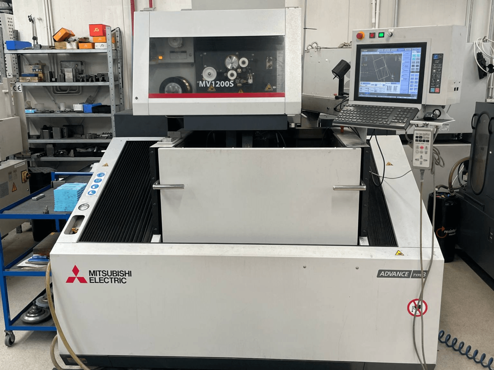 Mitsubishi MV1200S Tel EDM makinesinin önden görünümü; kontrol paneli ve çalışma alanı gösterilmekte olup, güvenlik uyarıları da görülebilmektedir.