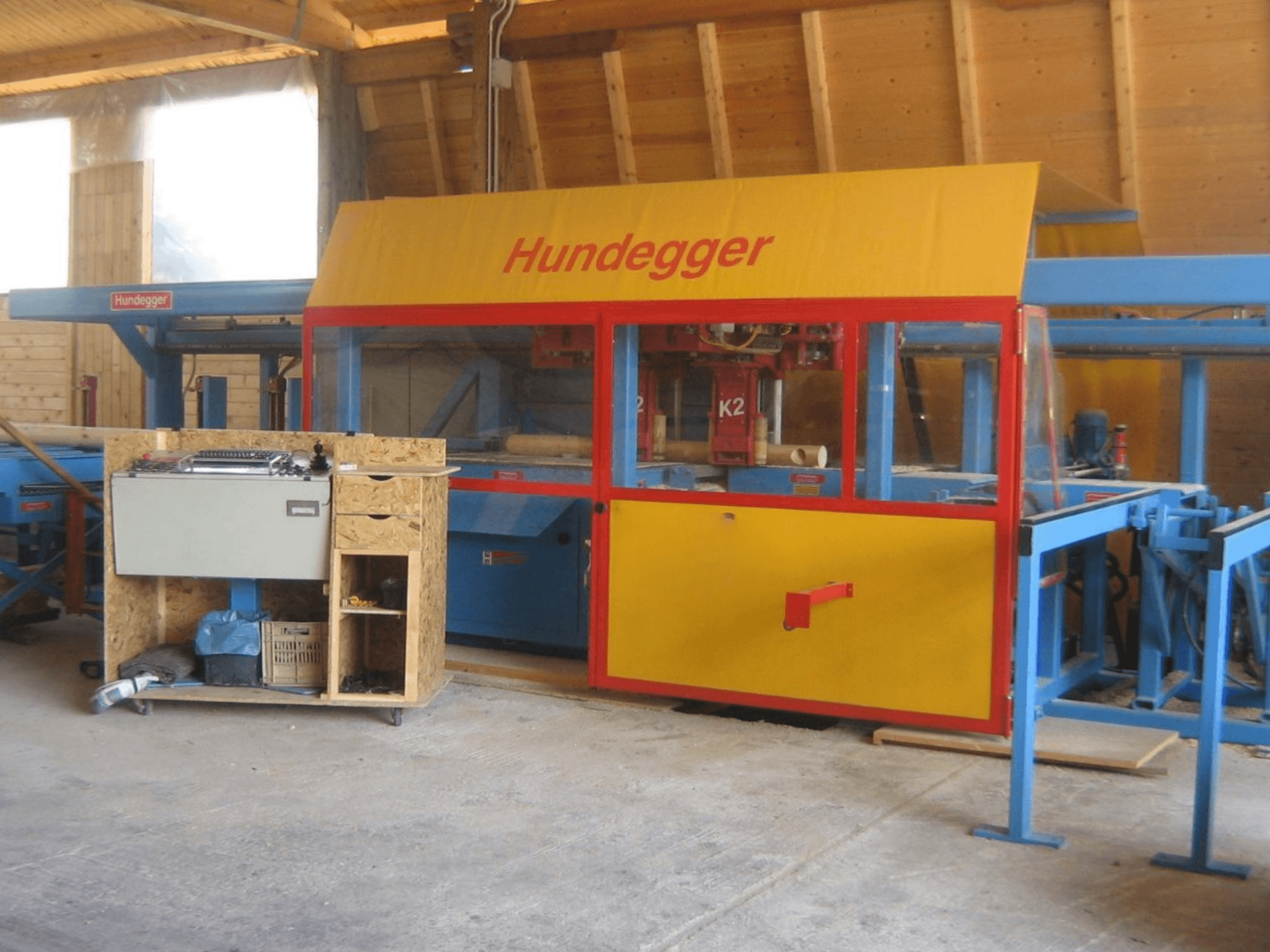 CNC Ahşap İşleme Merkezi HUNDEGGER K2 4 assi bir atölyede, çalışma tezgahı ile sarı ve mavi tasarımı gösteren önden görünüm.