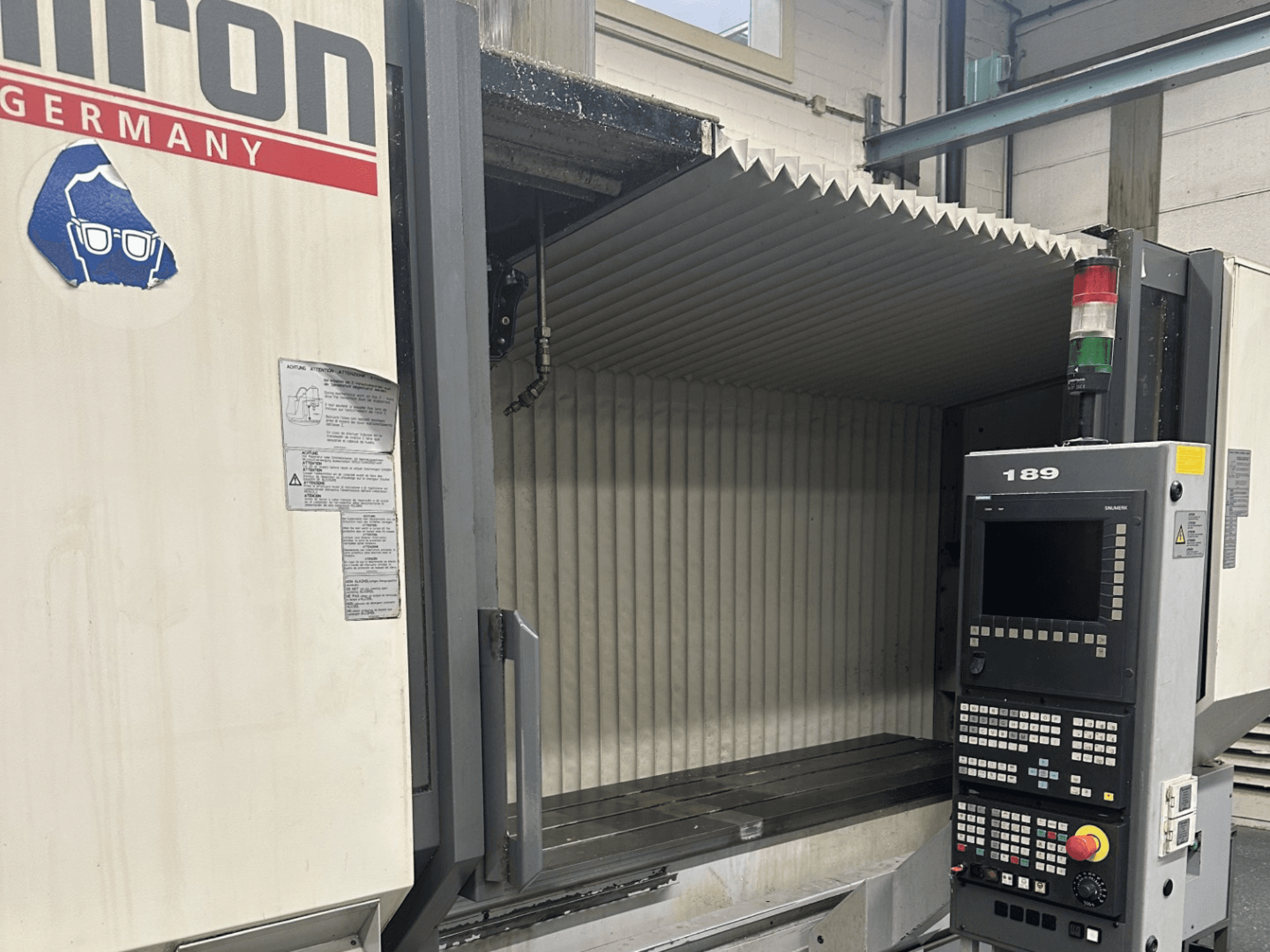 Alt metin: Açık bir çalışma alanına sahip, düğmeleri ve ekranı gösteren bir kontrol paneline sahip bir Hermle CNC makinesinin önden görünümü.