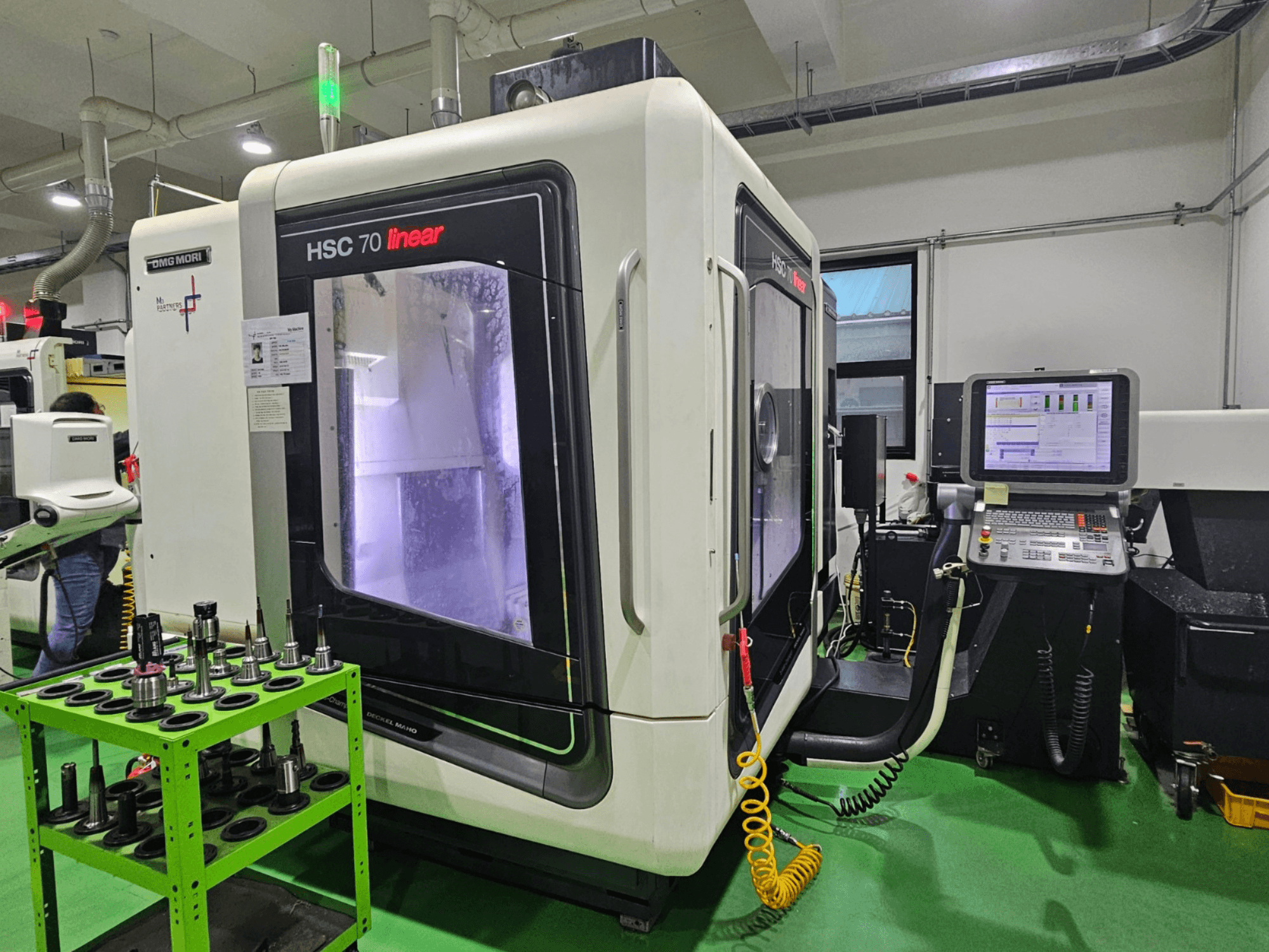 DMG MORI HSC 70 lineer üniversal işleme merkezi, önden görünüm, yeşil tabla üzerinde takım tutucular ve görünür bir kontrol paneli.