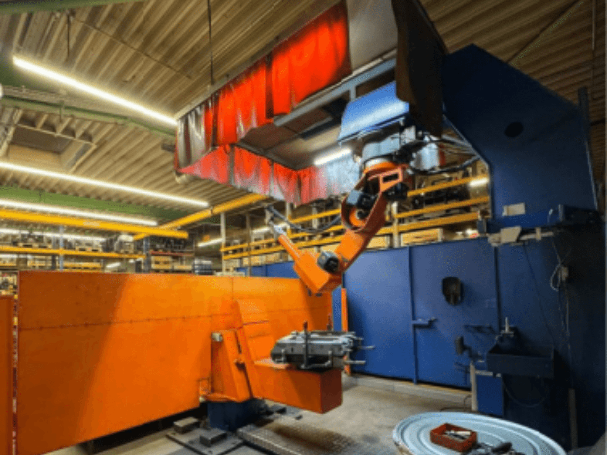 CLOOS Romat 350 e kaynak robotu endüstriyel bir ortamda, turuncu kollu önden görünüm, baş üstü perdesi altında bir iş parçasını tutuyor.