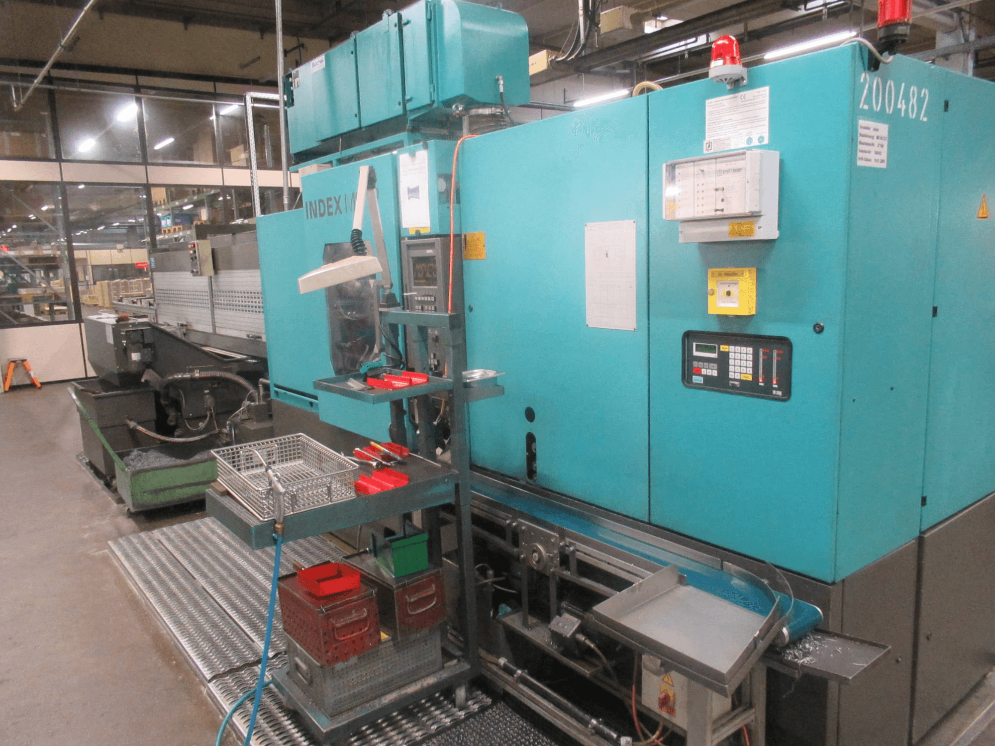 Fabrikadaki turkuaz INDEX makinesi, yandan görünüm; kontrol paneli, alet deposu ve konveyör bulunmaktadır. Görünür kutular, hortumlar ve alarmlar.