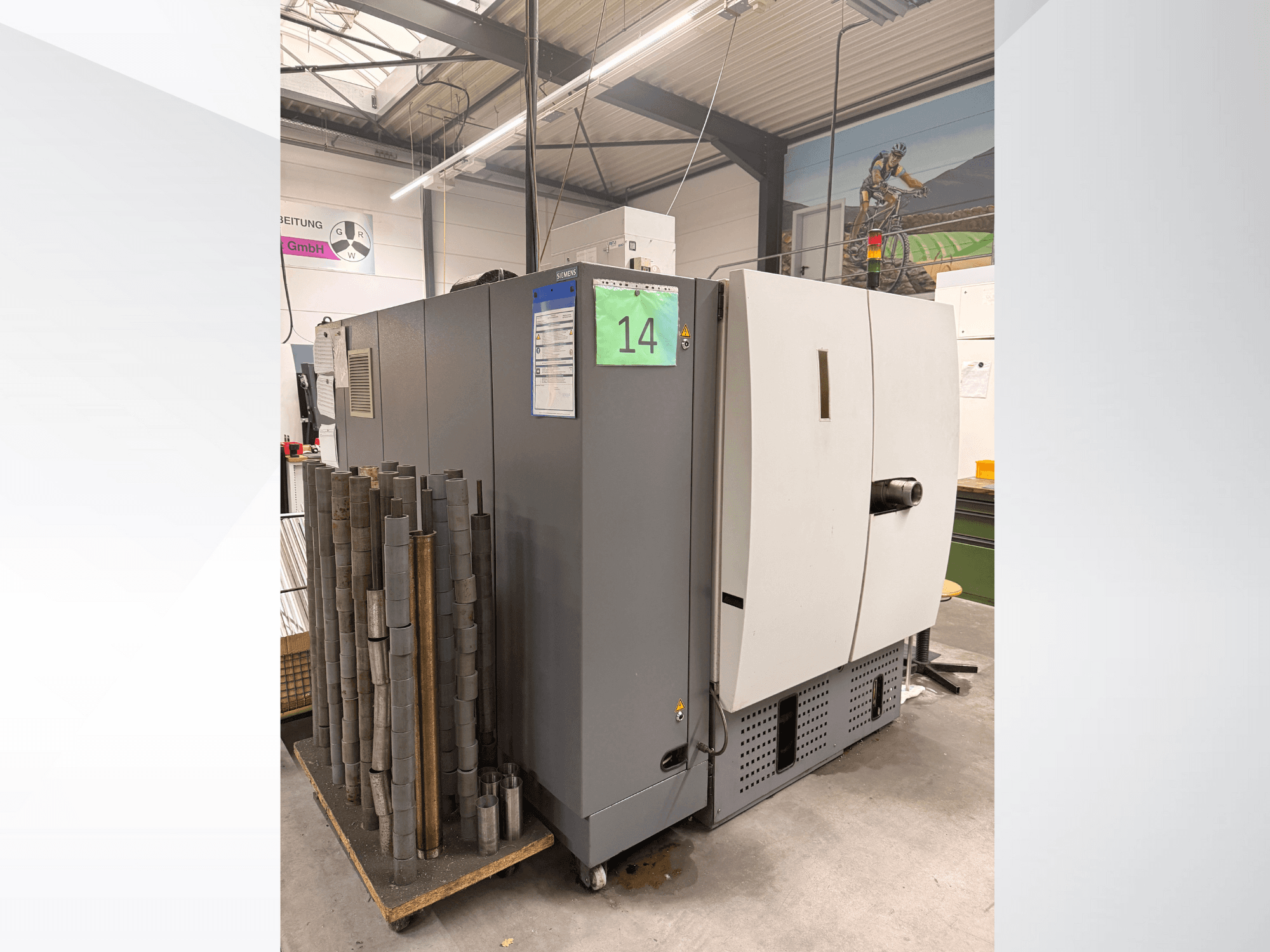 Siemens'ten gri endüstriyel makine, yandan görünüm, yanında silindirik metal parça stoğu ve ekipman etiketleri görünüyor.