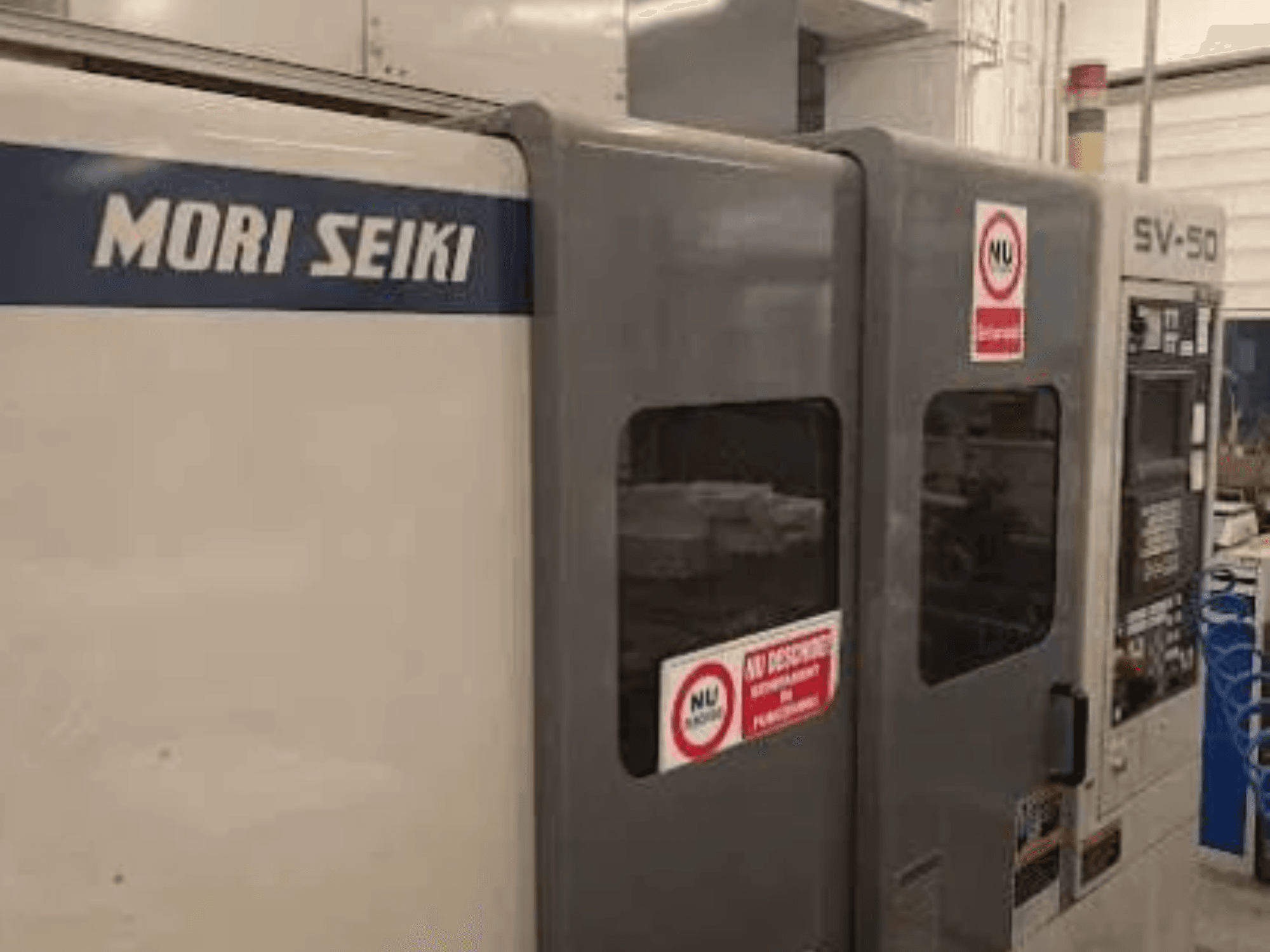 MORI SEIKI SV-50 CNC makinesi, yandan görünüm; kontrol paneli ve güvenlik uyarı etiketleri bulunmaktadır.