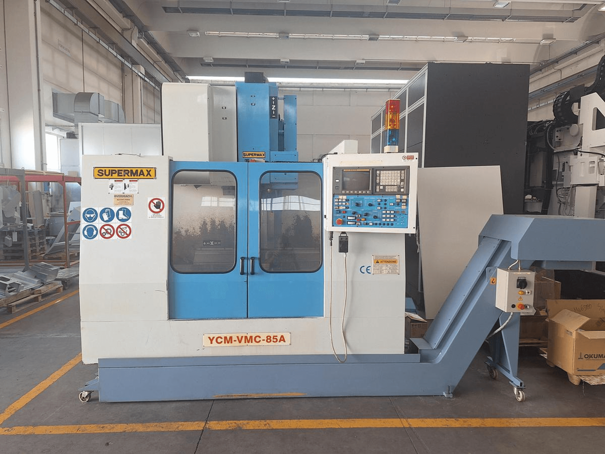 Kontrol paneli ve güvenlik etiketleriyle donatılmış bir fabrikada bulunan SUPERMAX YCM-VMC-85A dikey işleme merkezi, önden görünüm.