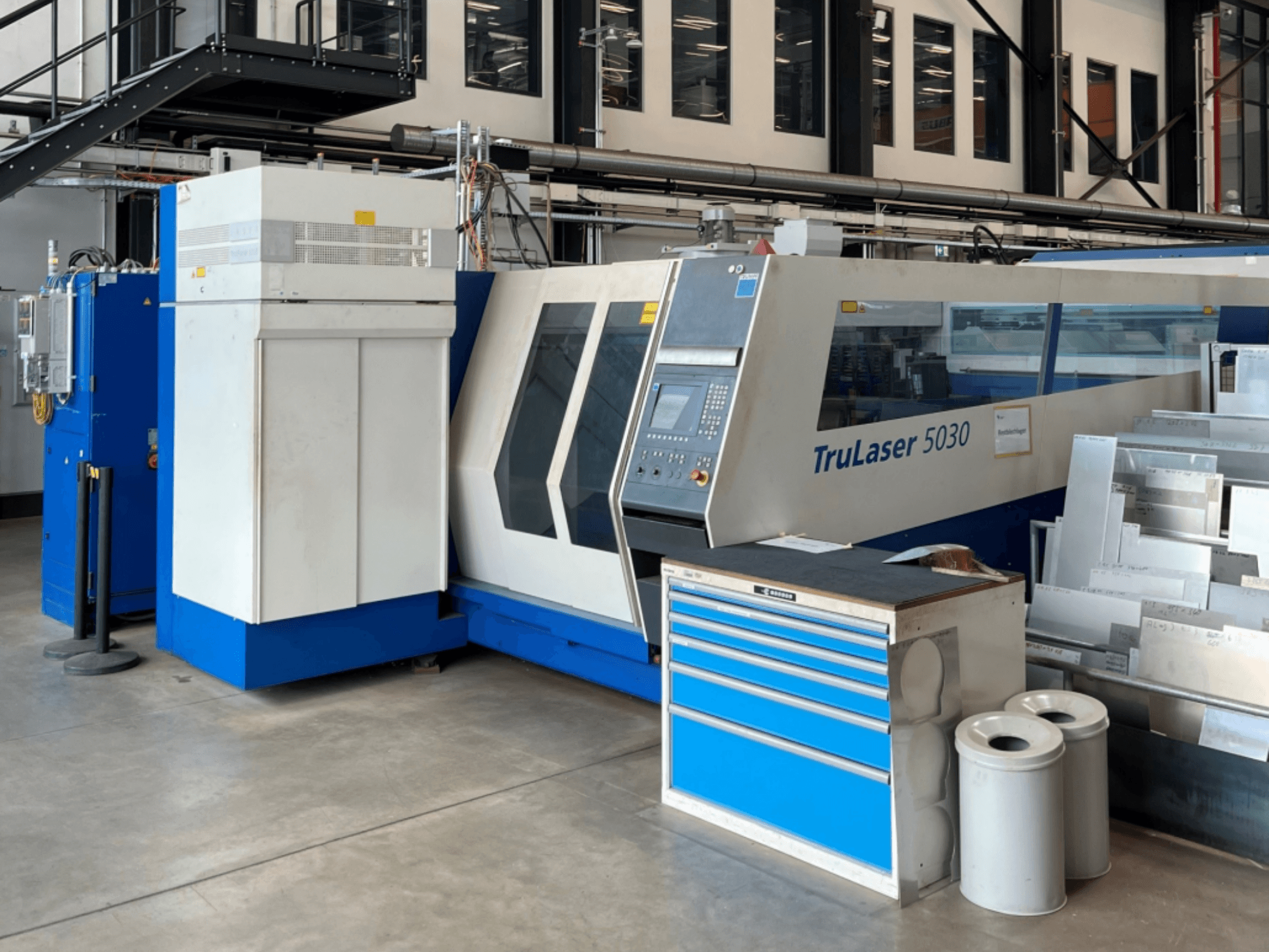 TRUMPF TruLaser 5030 CO₂ lazer kesim makinesinin önden görünümü; kontrol paneli ve yanında metal levhaların bulunduğu çalışma tezgahı görülmektedir.