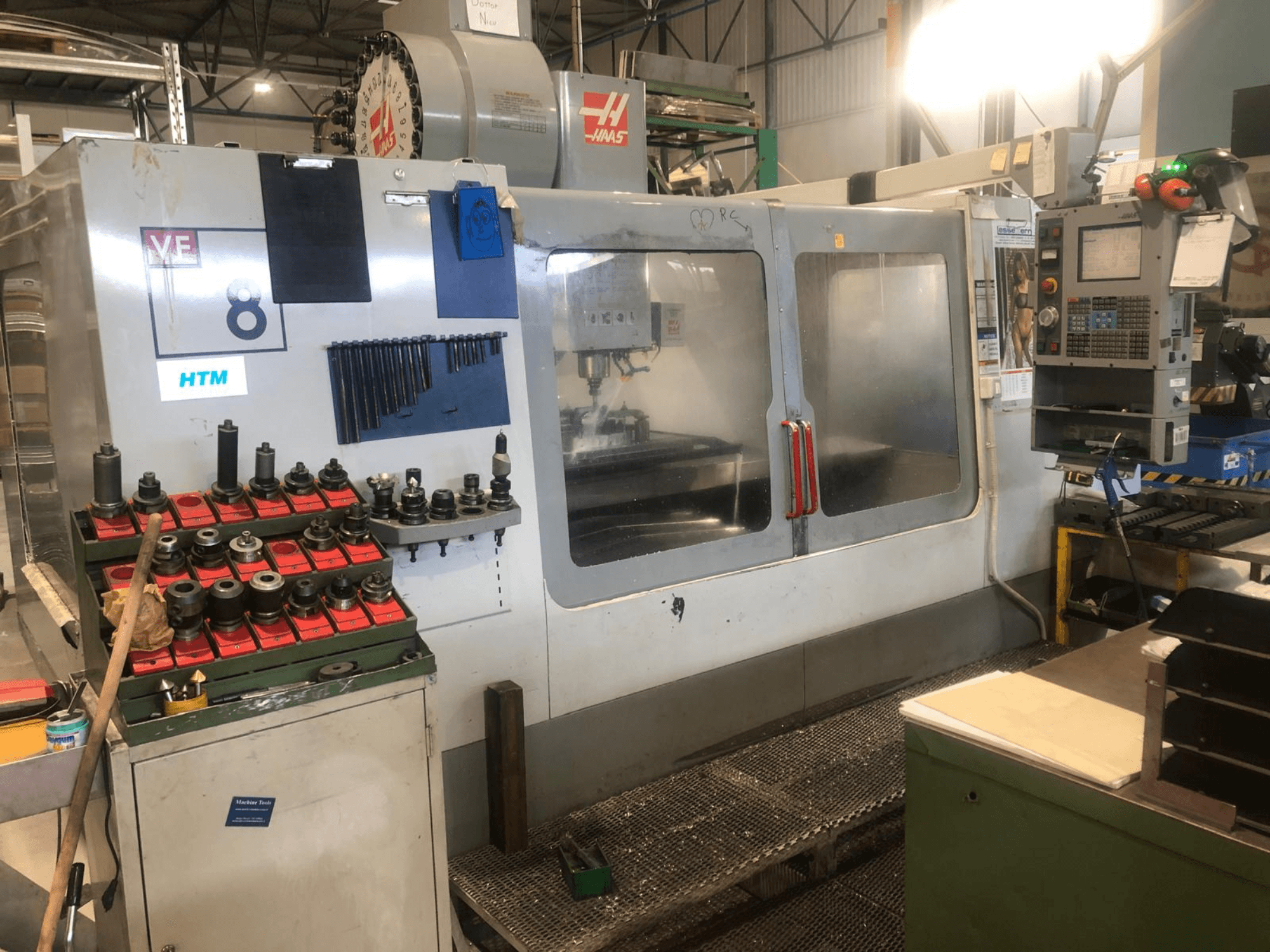 Atölyedeki Haas CNC makinesi, önden görünüm; görünür model detayları ve düzenli takım tutucuları ile takımları ve kontrolleri gösterir.