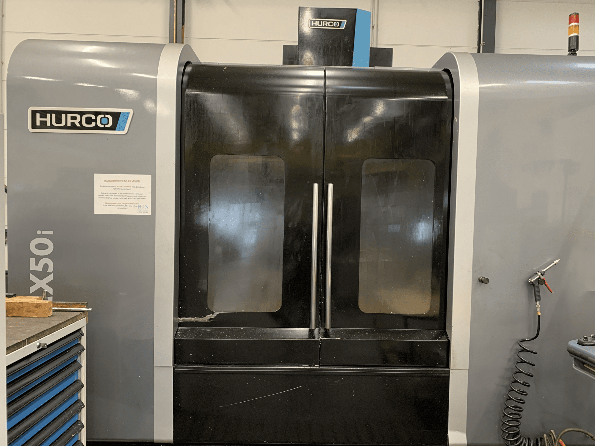 Çift kapılı bir Hurco VX50i CNC makinesinin önden görünümü, teknik kontrolleri ve yan taraftaki bakım işaretini göstermektedir.