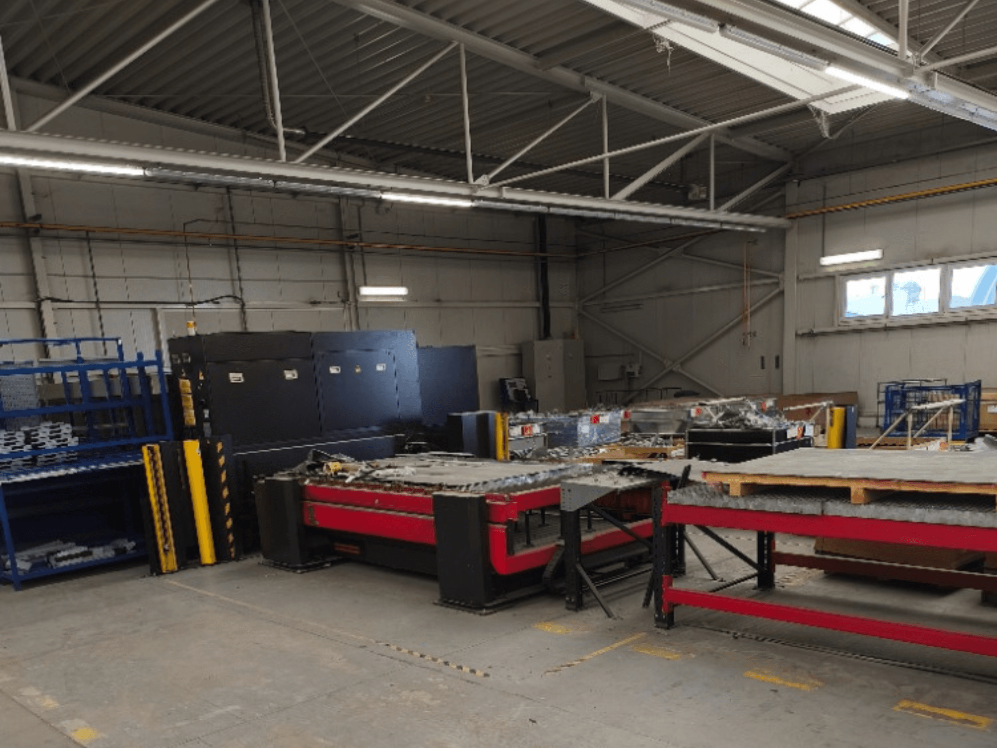 Bir atölyede bulunan AMADA LCG 3015 AJ fiber lazer kesim makinesi; makine önden görülürken arka planda depolama rafları ve malzemeler yer almaktadır.