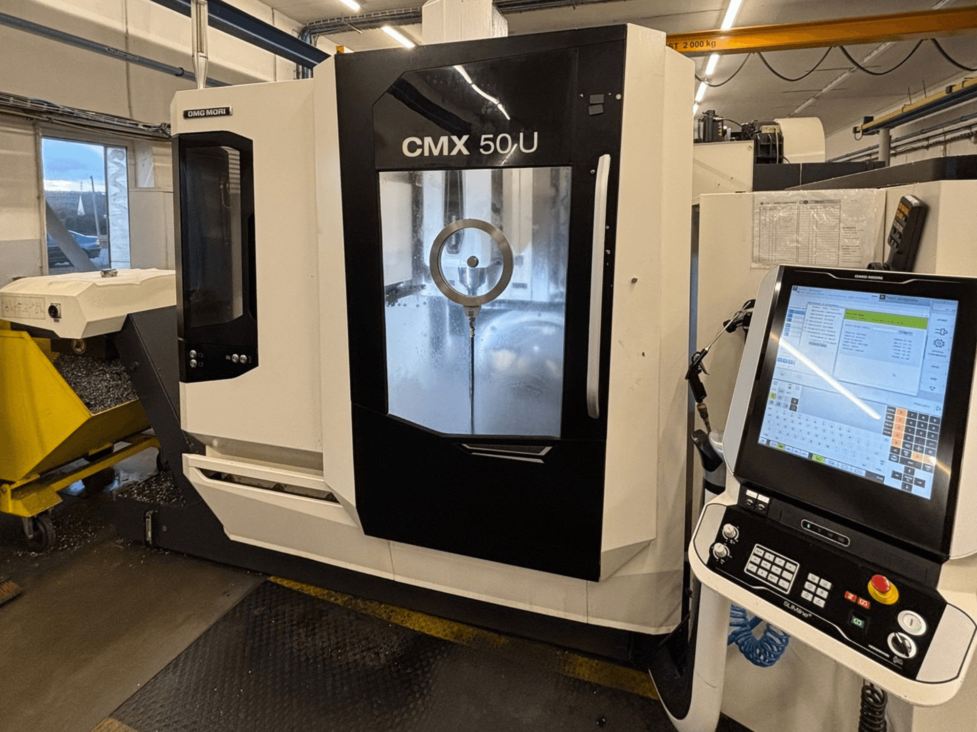 DMG MORI CMX 50 U dikey işleme merkezi, kontrol paneli ve cam ekran ile önden görünüm, iç mekaniği gösterir.