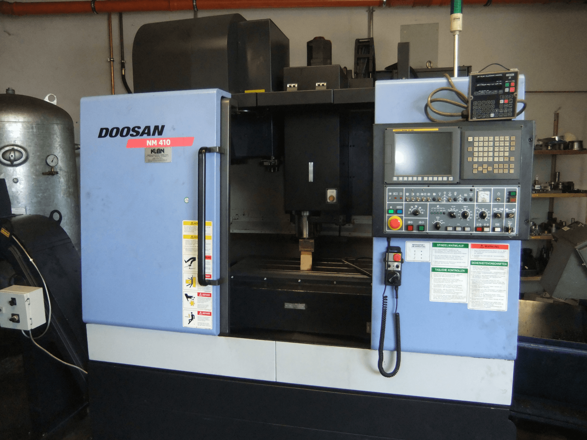 DOOSAN NM 410 dikey işleme merkezi, önden görünüm; kontrol paneli, iş mili bölgesi ve güvenlik etiketleri gösterilmektedir.