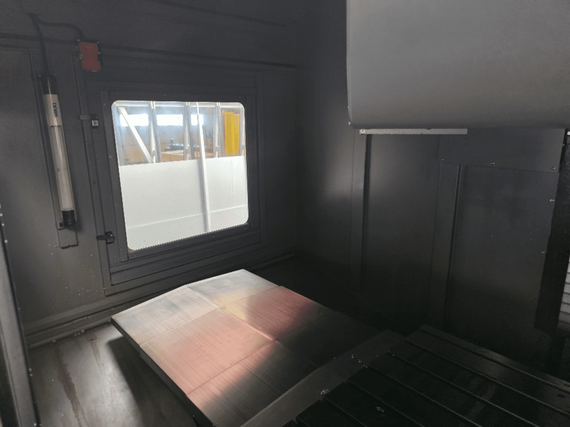 Cilalı metal plakalı, yan pencereli ve minimum aydınlatmalı bir makine muhafazasının iç görünümü. Marka görünmüyor.