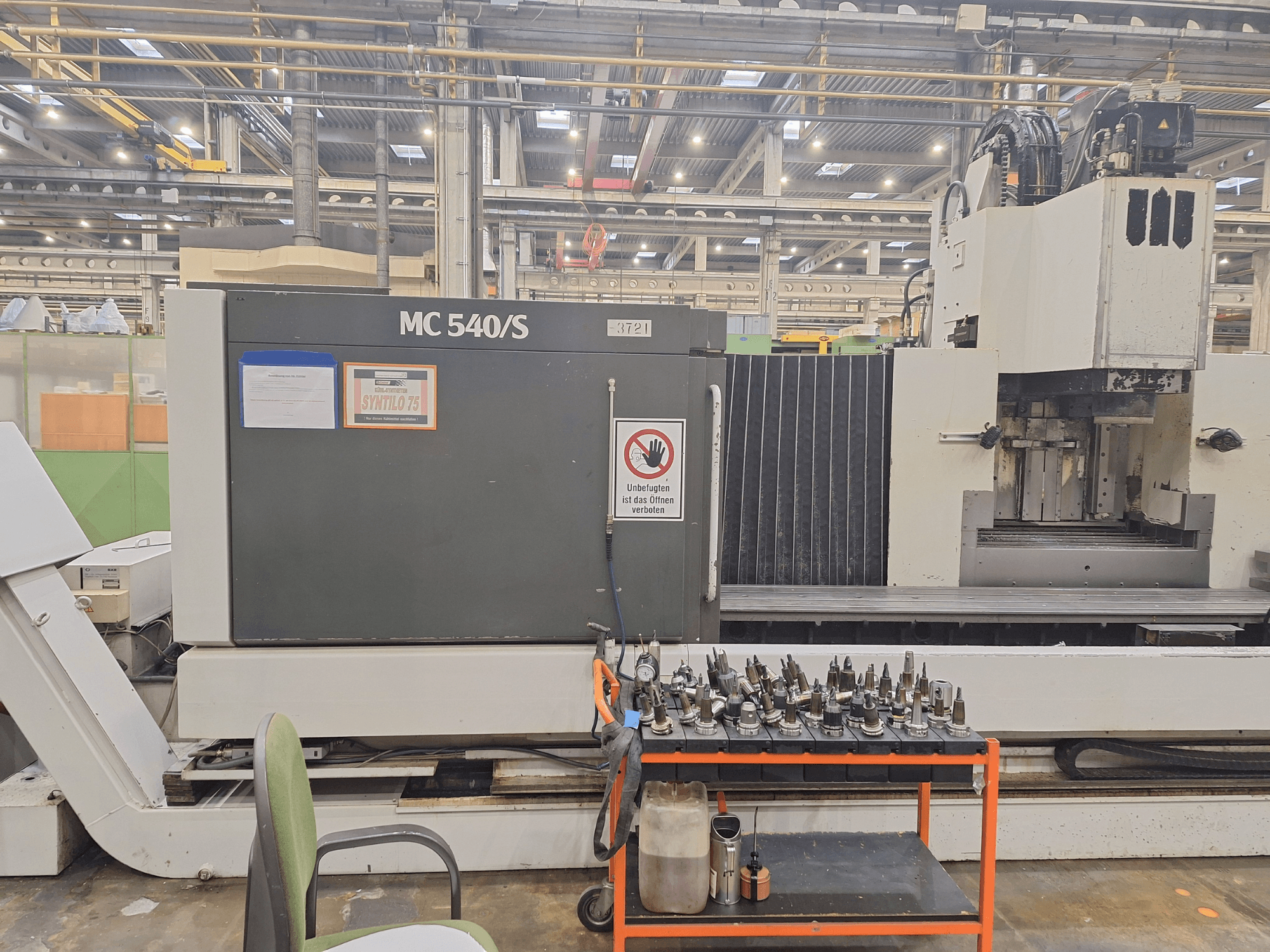 Dikey işleme merkezi STAMA MC 540S bir atölyede, yan tarafta bir araba üzerinde takım tutucular ile yandan görünüm.