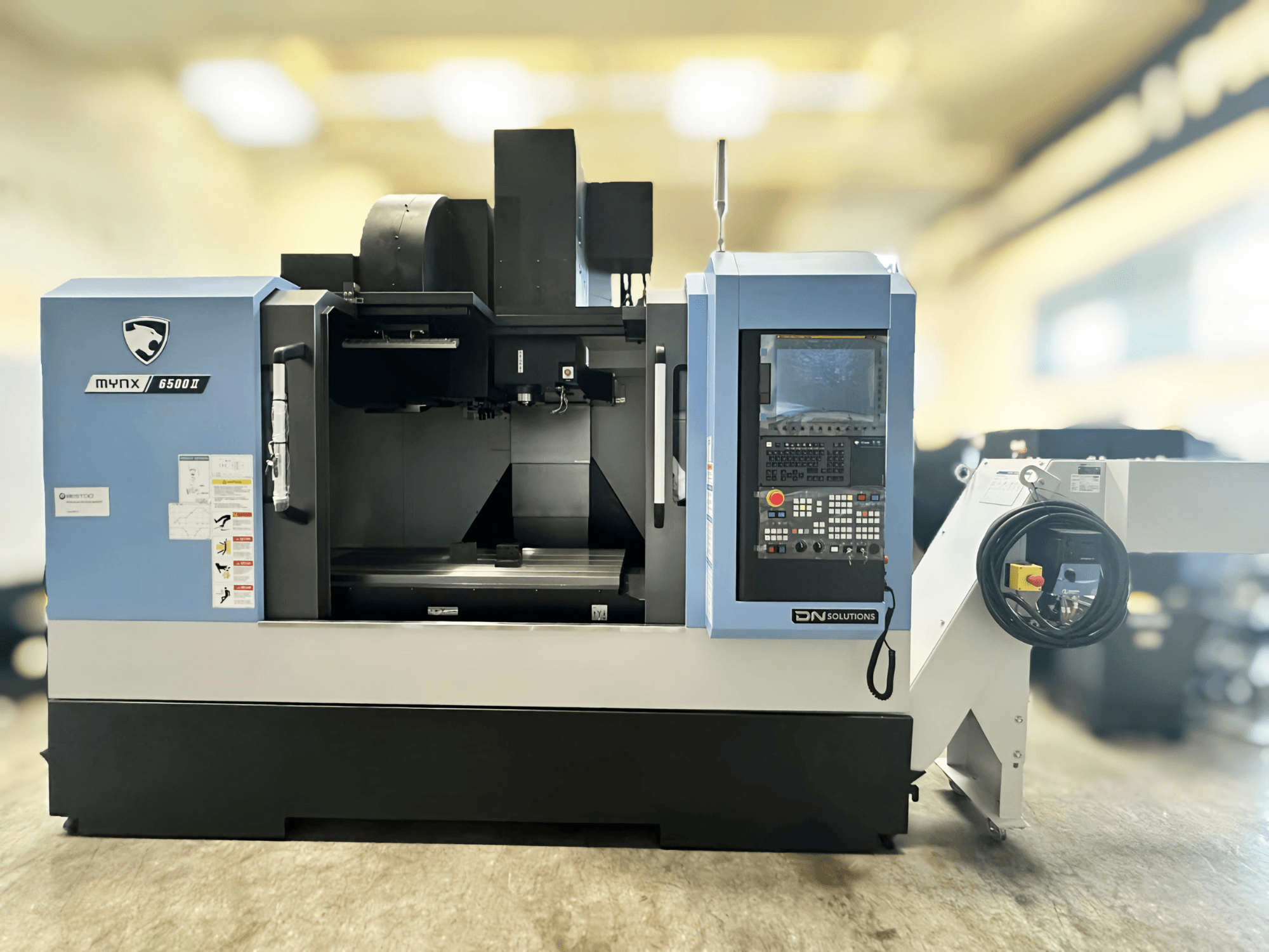 Dikey işleme merkezi DN SOLUTIONS MYNX 6500 II, önden görünüm, kontrol paneli ve açık işleme alanı.