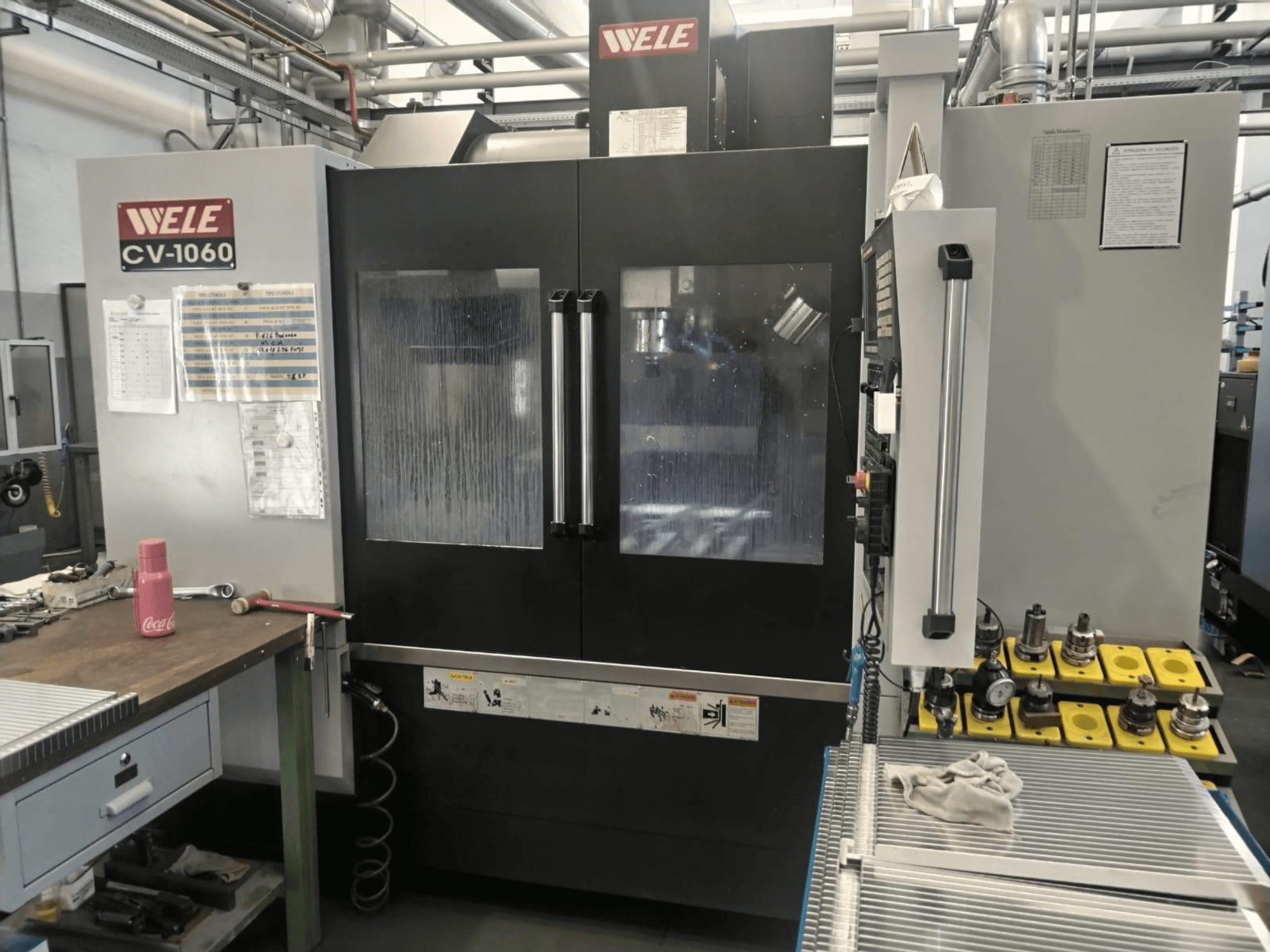 Wele CV-1060 dikey işleme merkezi, önden görünüm, kontrol paneli ve görünür çalışma alanı ile birlikte; aksesuar aletleri ve güvenlik etiketleri dahildir.