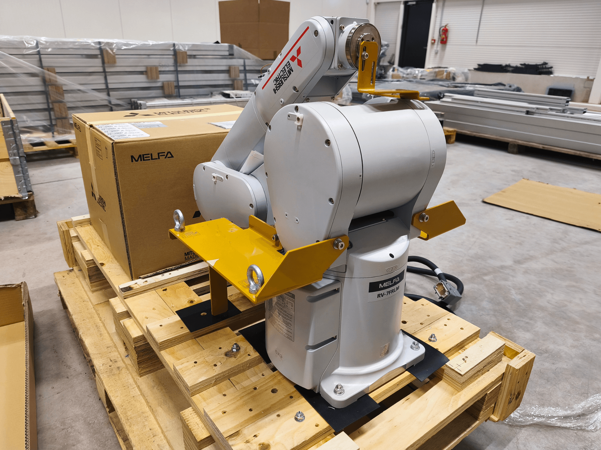 MELFA RV-7FLM endüstriyel robot kolu ahşap palet üzerinde, önden görünüm, sarı destek braketleri ve gri ambalaj kutusu ile.